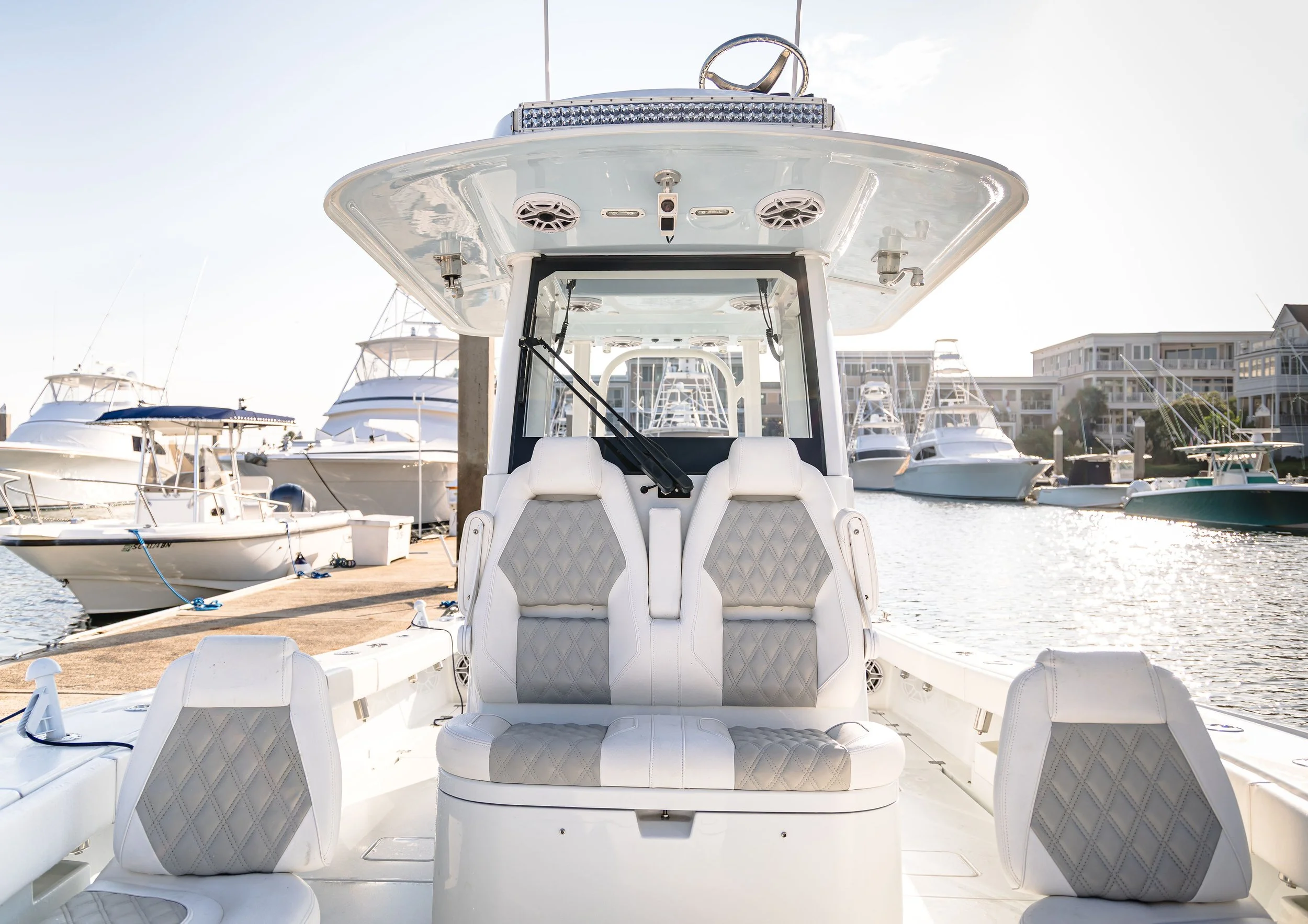 View of a luxury boat docked at a marina, showing the boat's seating area with white leather cushions, surrounded by other boats and modern houses in the background.
