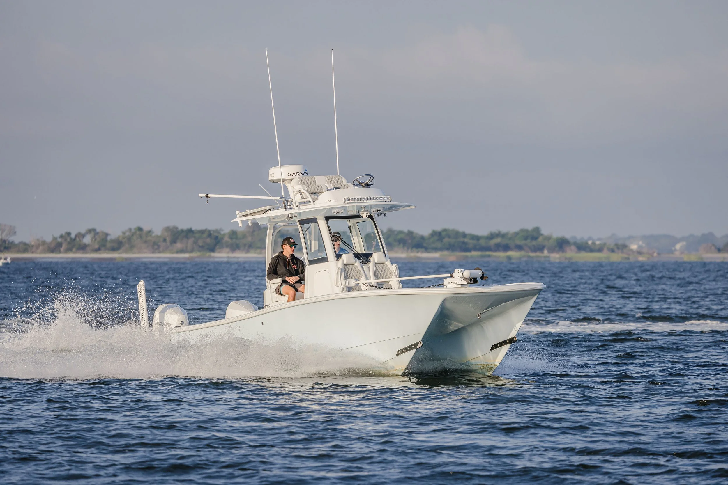 A white motorboat with two occupants, sailing on water with a shoreline in the background, and cloudy sky overhead.