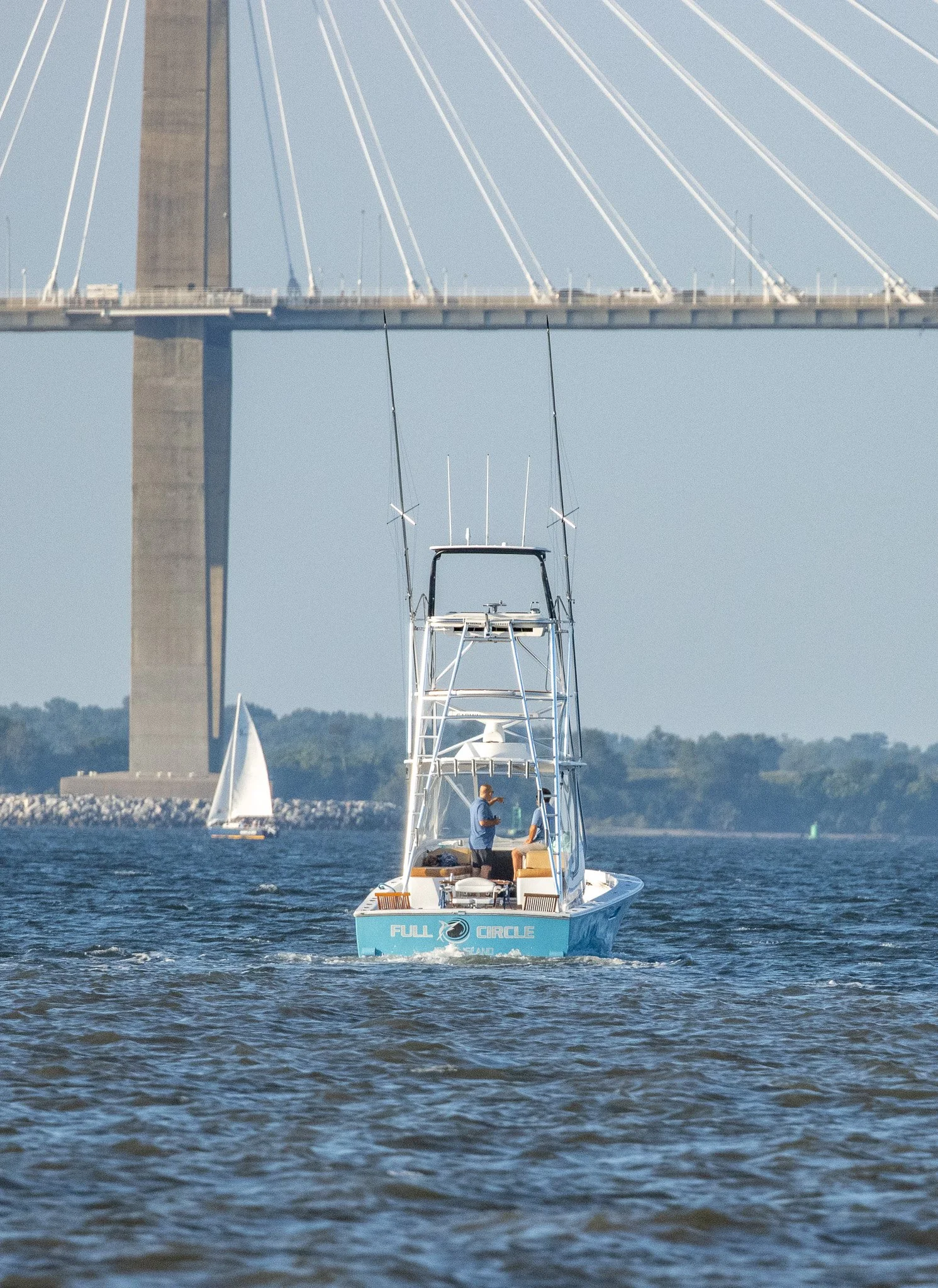 A motorboat named 'Full Circle' cruising on the water under a large bridge with suspension cables, a sailboat in the background, and two people on the boat.
