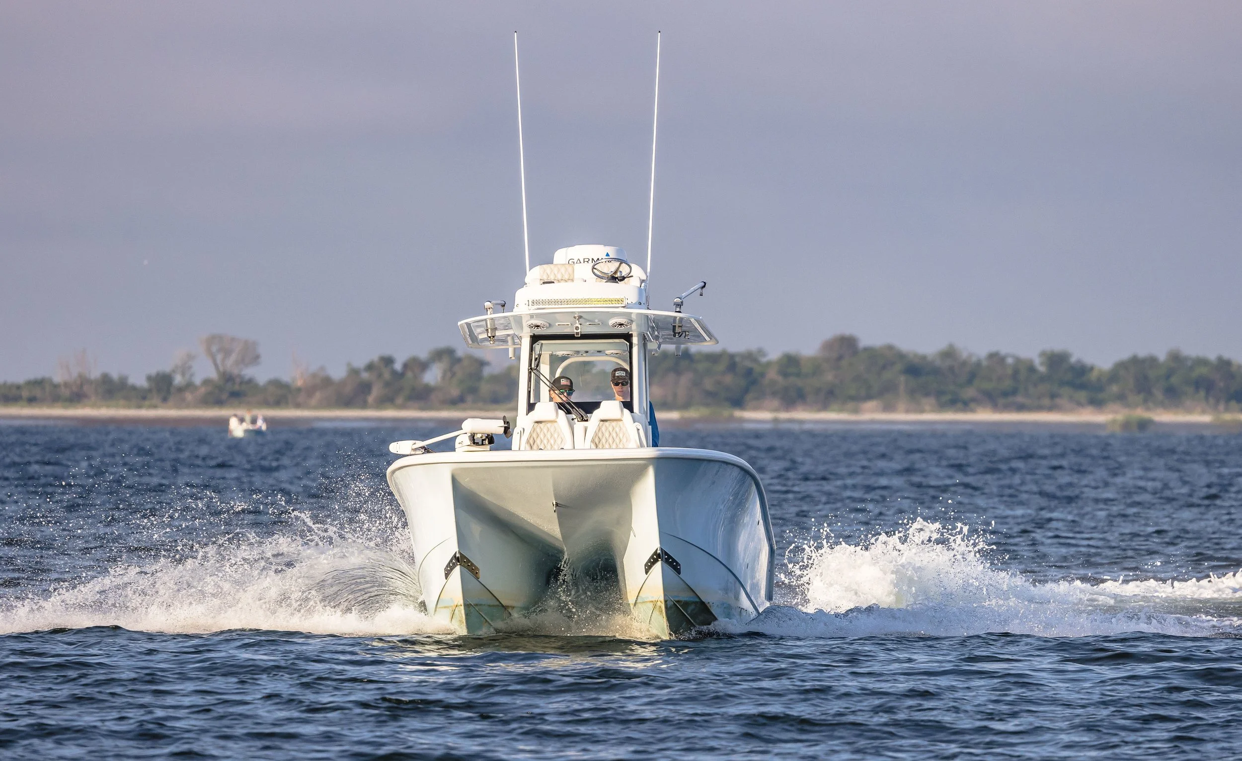 A white motorboat moving through a body of water, with two people onboard wearing sunglasses. The boat is creating a wake as it speeds forward, with a distant shoreline and trees in the background under a partly cloudy sky.