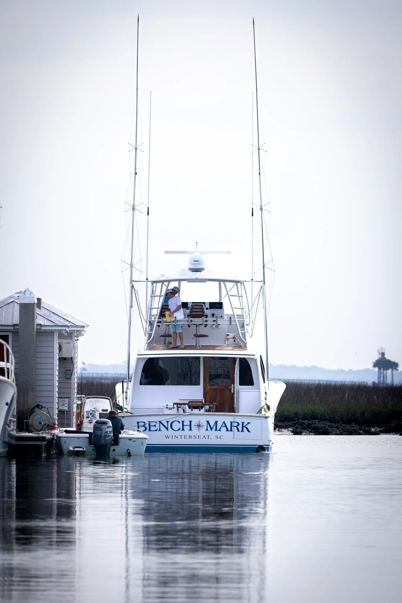 A white yacht named 'Bench Mark' docked at a marina with two men on the upper deck, surrounded by calm water and marshland in the background.
