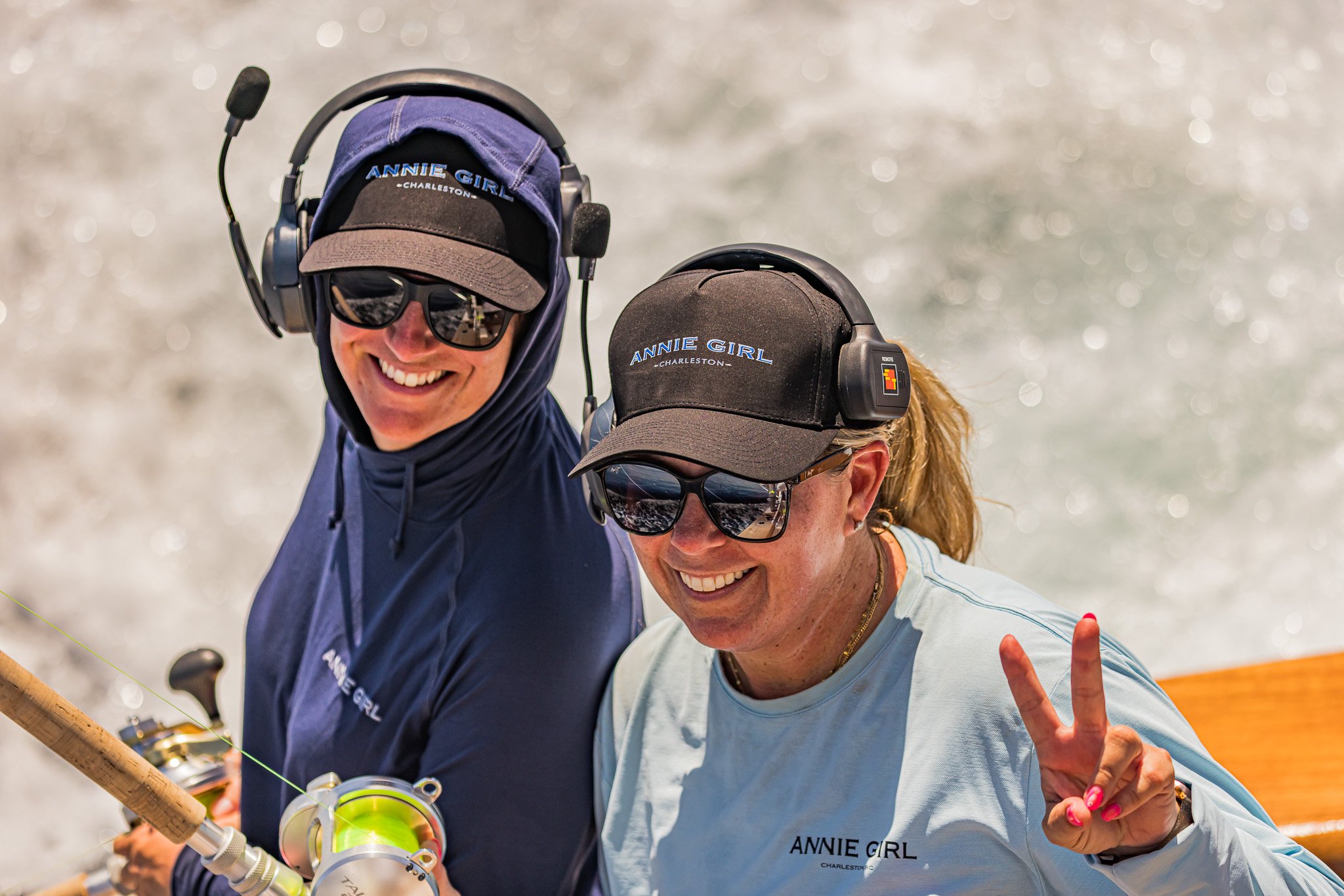 Two women smiling, wearing headsets and hats with 'Annie Girl Charleston' logo, posing for a photo on a boat or near water.