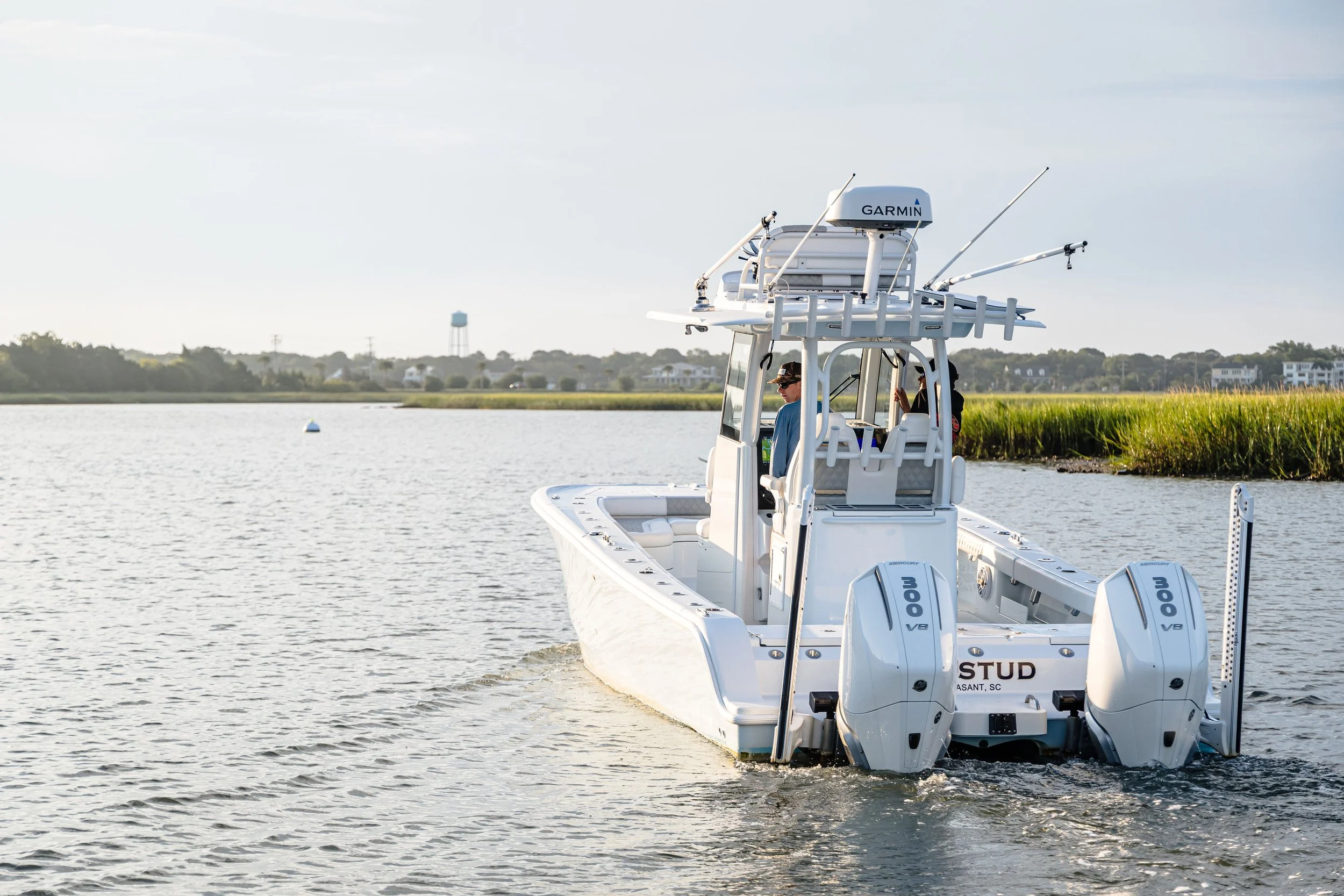 A white fishing boat with dual outboard motors on a body of water. Two people are on the boat, one wearing a blue shirt and sunglasses, and the other partially visible. The background includes tall grass, distant houses, and a water tower, under a pa