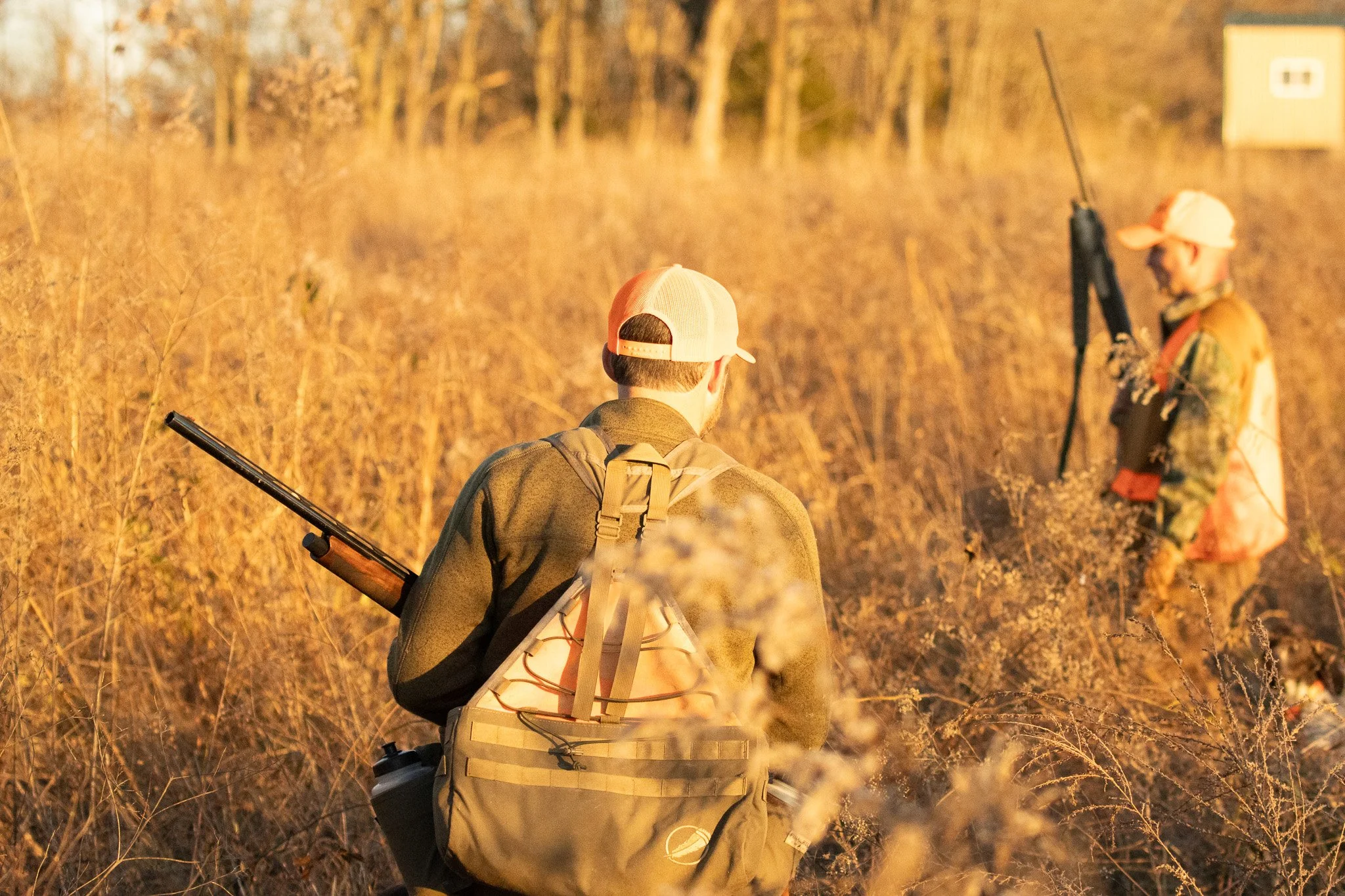 Two people walking through a field at sunset, carrying rifles and wearing backpacks and hats.