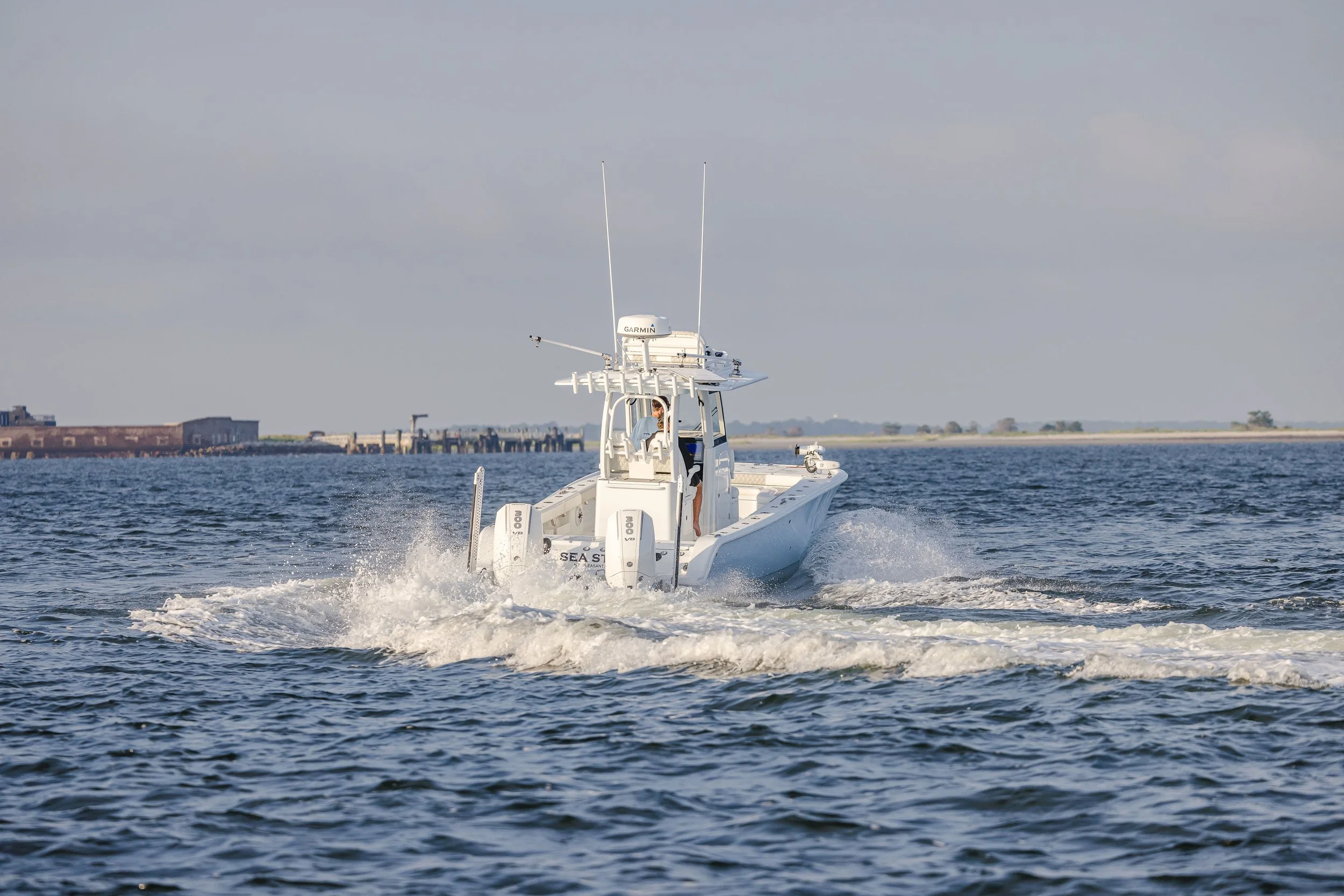 A white motorboat speeding across the water with a Garmin radar and antennas on top, leaving a wake behind it, with a shoreline and cloudy sky in the background.