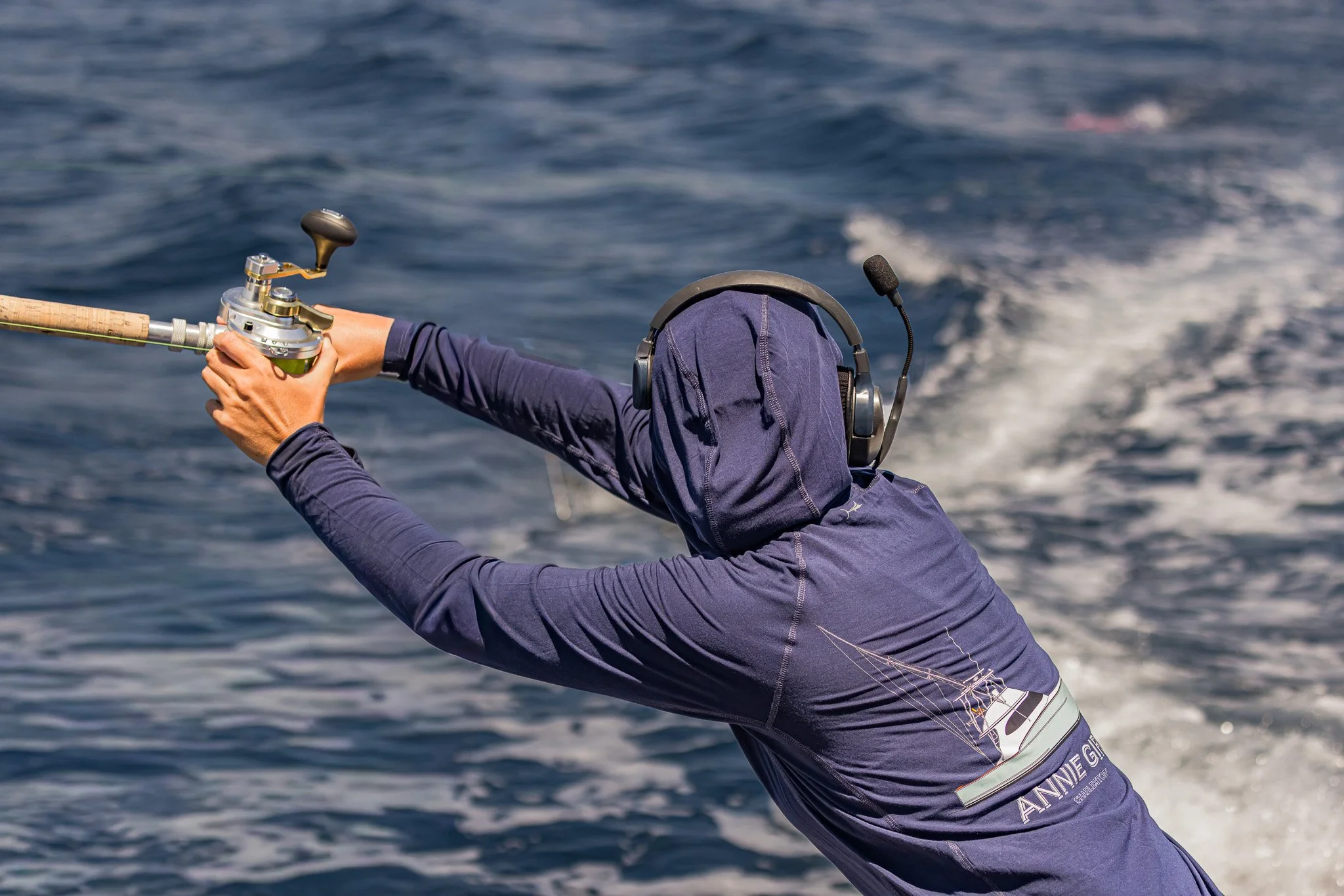 A person wearing a navy jacket and a head covering, holding a fishing rod over the water while fishing on a boat.