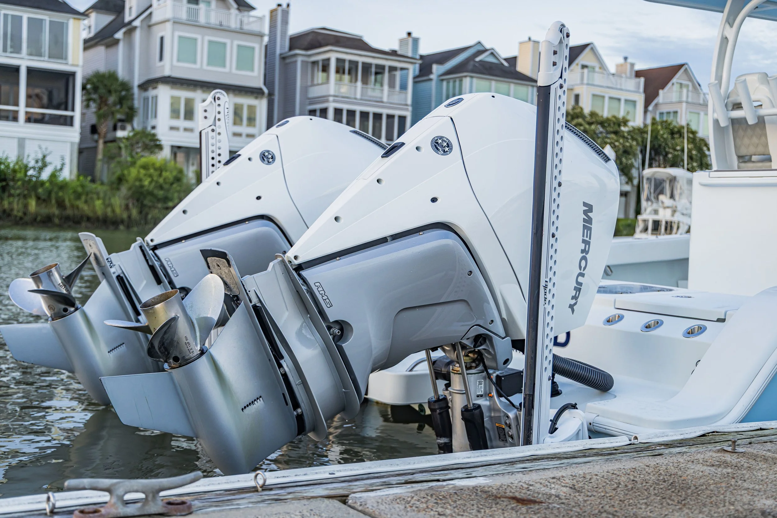 Close-up of a white boat engine with several propellers, docked at a marina with residential buildings in the background.