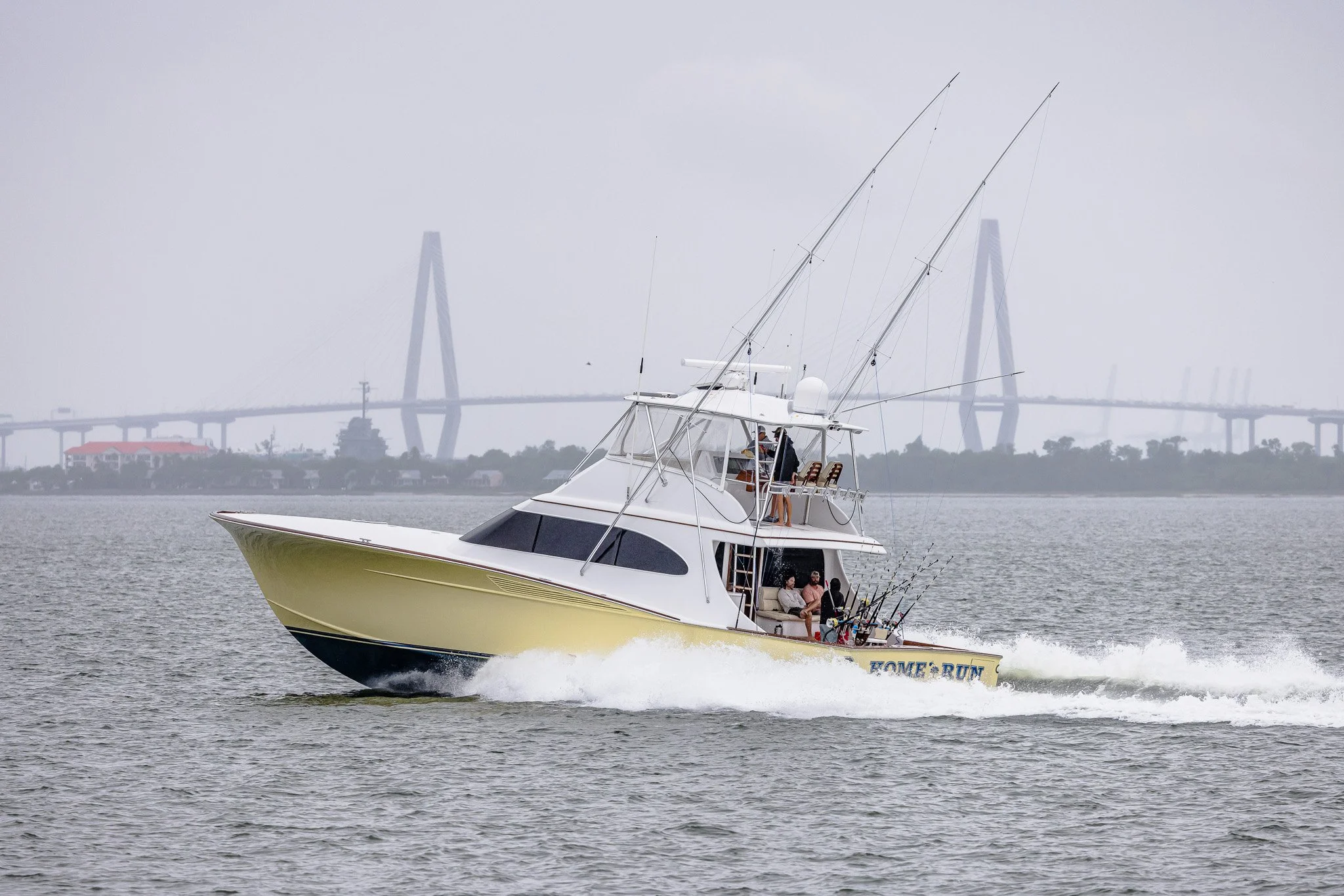 A motorboat with people on board speeding across the water, with fishing rods visible, and a city skyline with a bridge in the background.