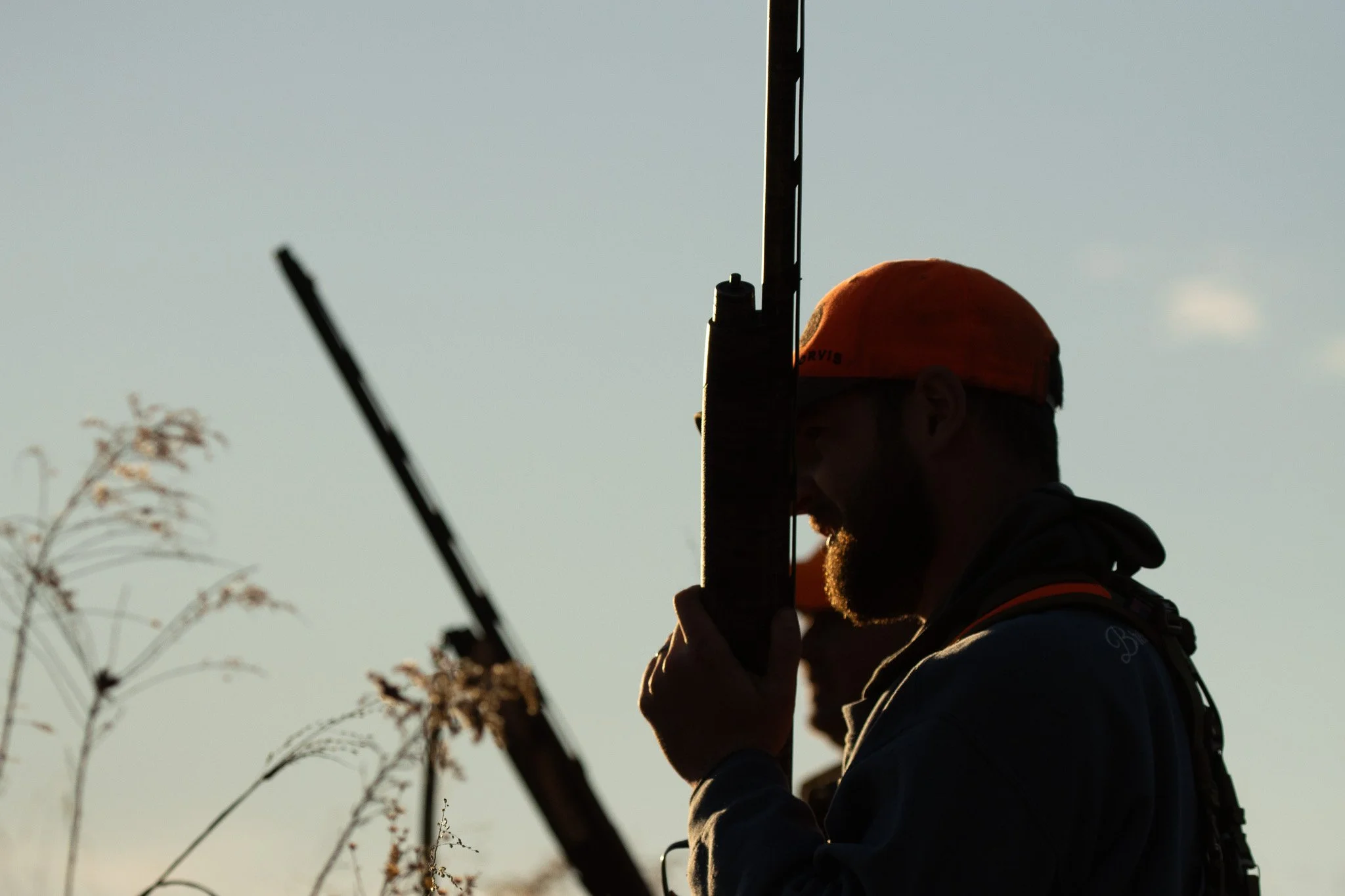 A person wearing an orange cap and dark clothing, seen in silhouette, is holding a smartphone or similar device, with the sky in the background.