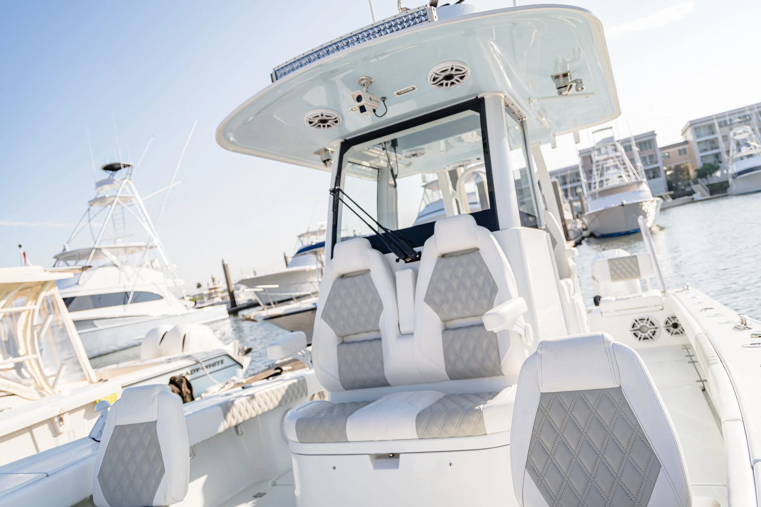 A close-up of a white fishing or recreational boat docked at a marina with other boats in the background and positioned against a backdrop of water and multi-story residential buildings.