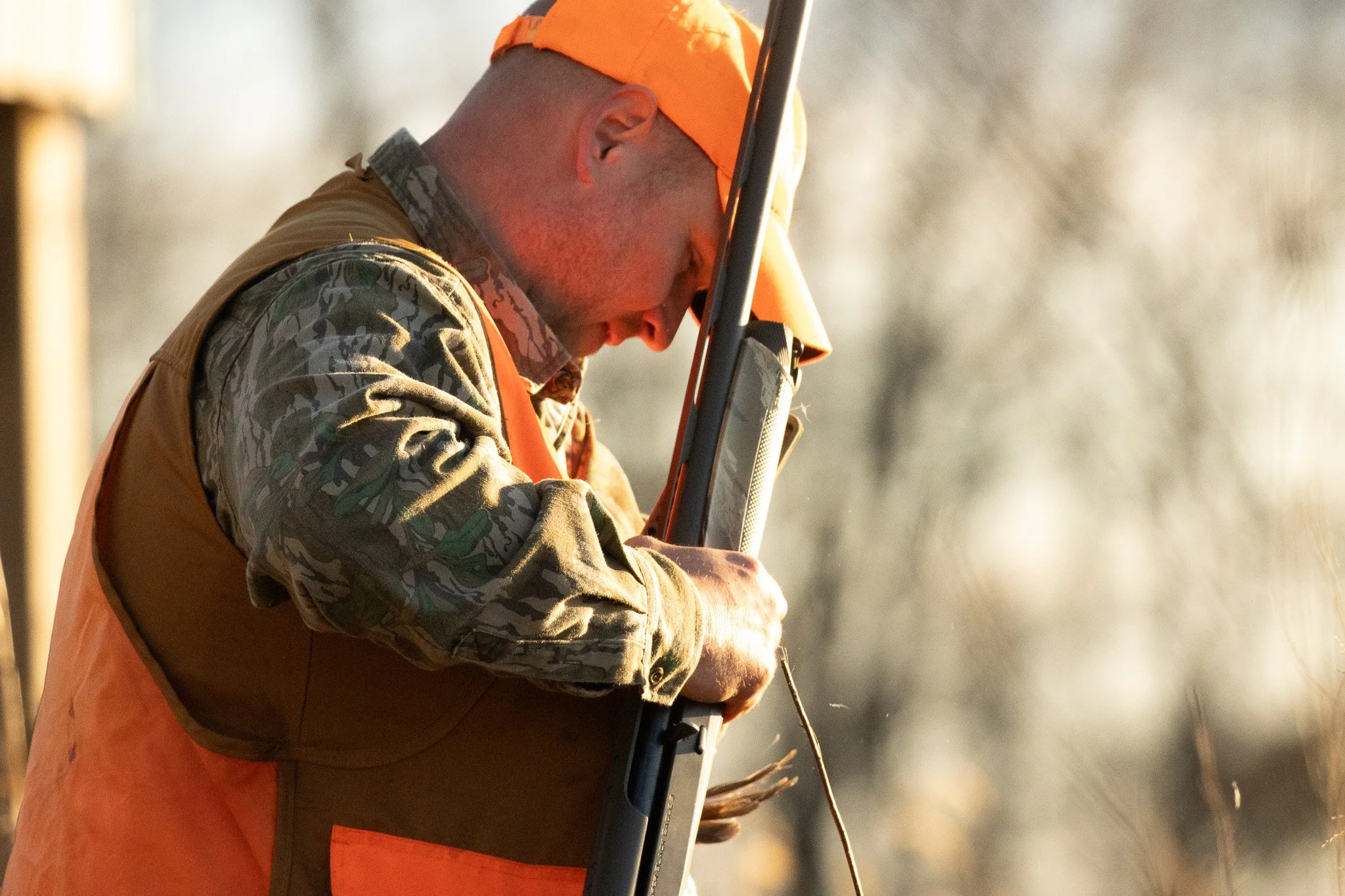 A person dressed in camouflage clothing and an orange safety vest and cap is holding and aiming a shotgun, with a focus on the person's head and upper body against a blurred outdoor background.