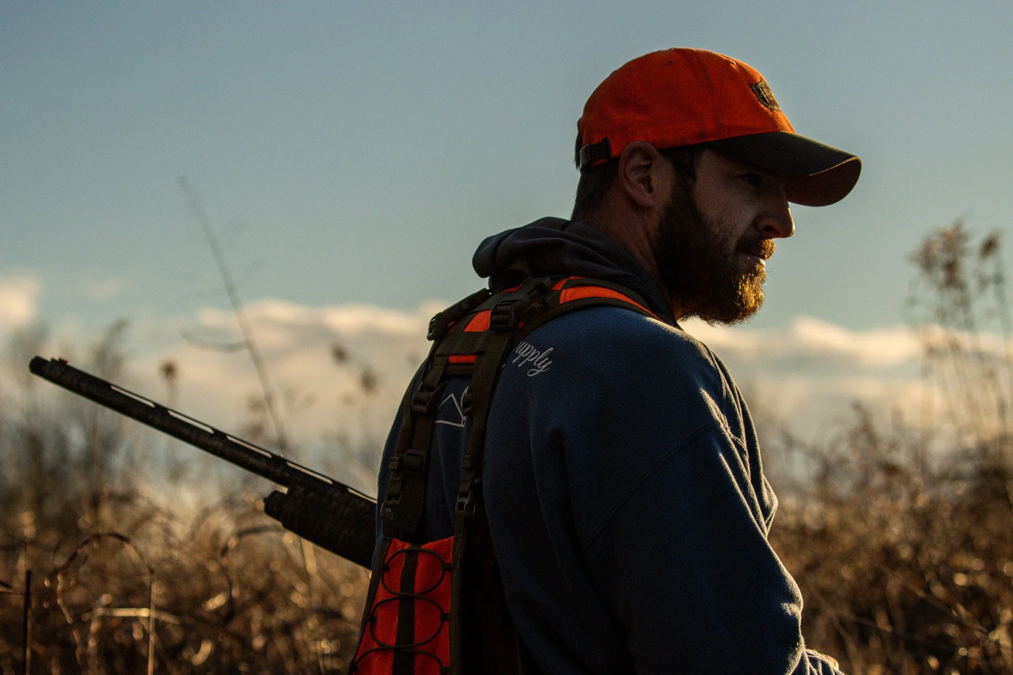 A man with a beard wearing an orange hat and dark jacket, standing outdoors during sunset, carrying a backpack and holding a walking stick.