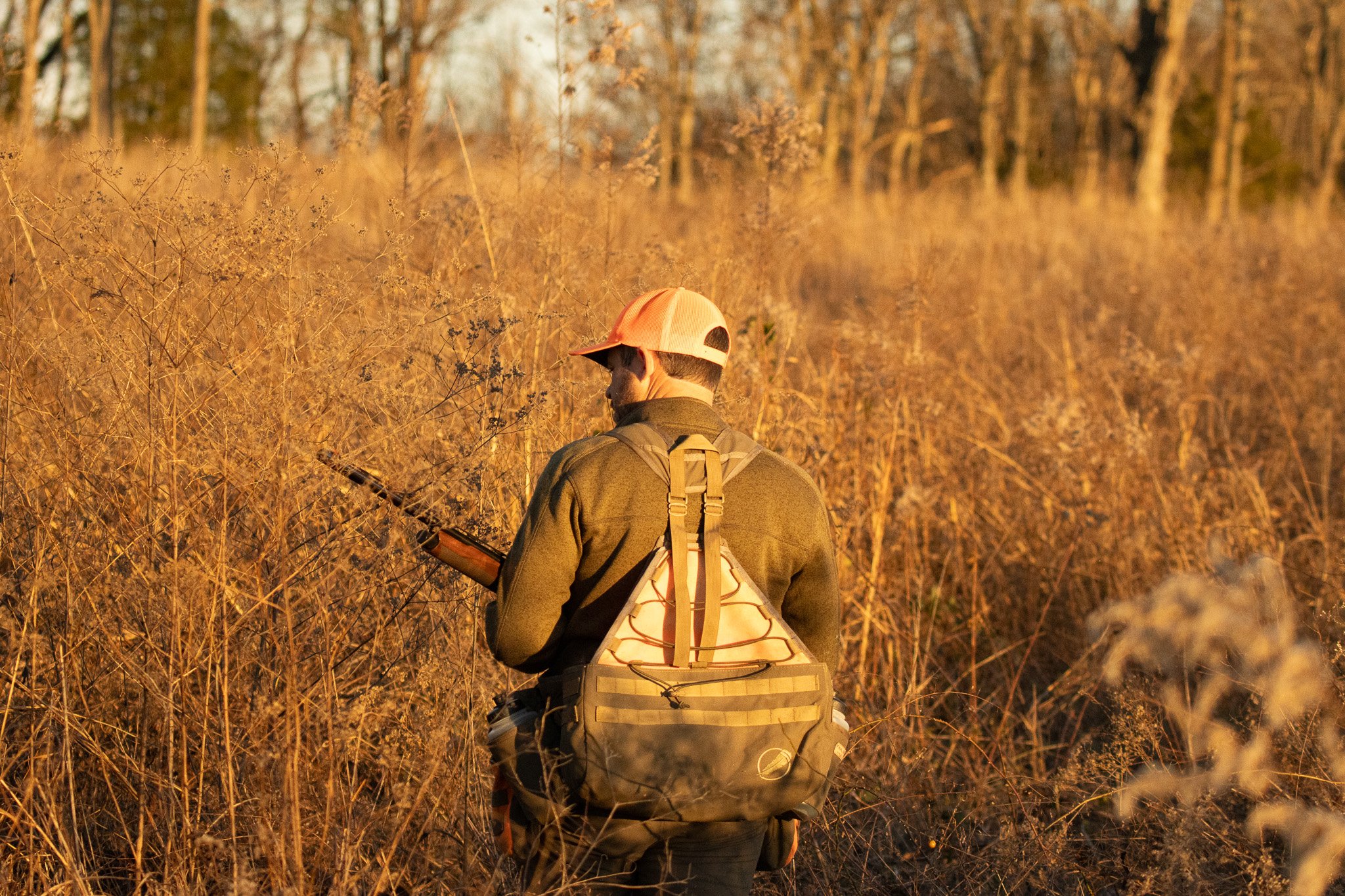 A person wearing a beige baseball cap, a green jacket, and a backpack, standing in a dry, grassy field with tall trees in the background, holding a tablet or notebook