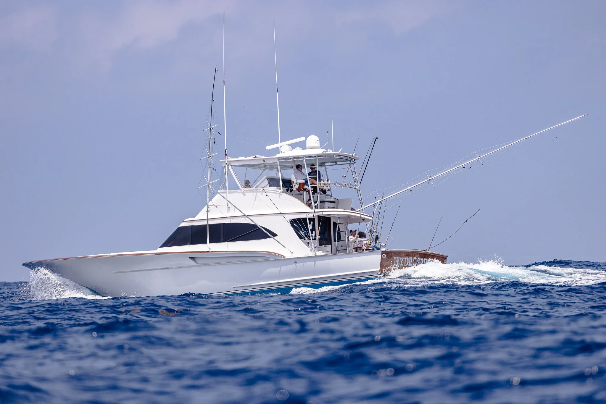 A white yacht with a brown hull named HydroSpear is sailing on the ocean with people on deck, fishing rods extended over the water, and a light blue sky above.