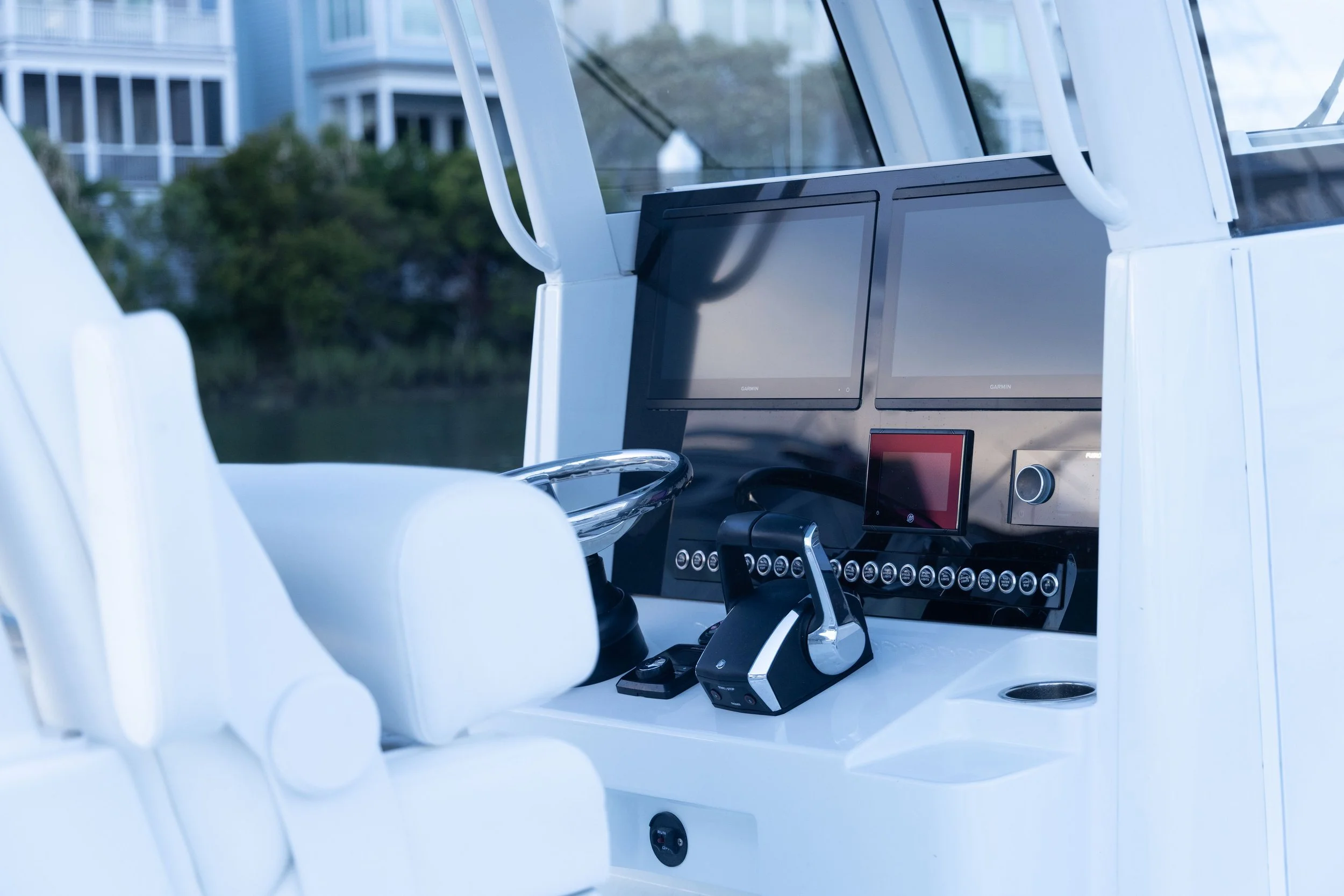 Interior of a boat cockpit with two Garmin navigational screens, a steering wheel, and various control buttons, with a view of houses and trees outside the window.