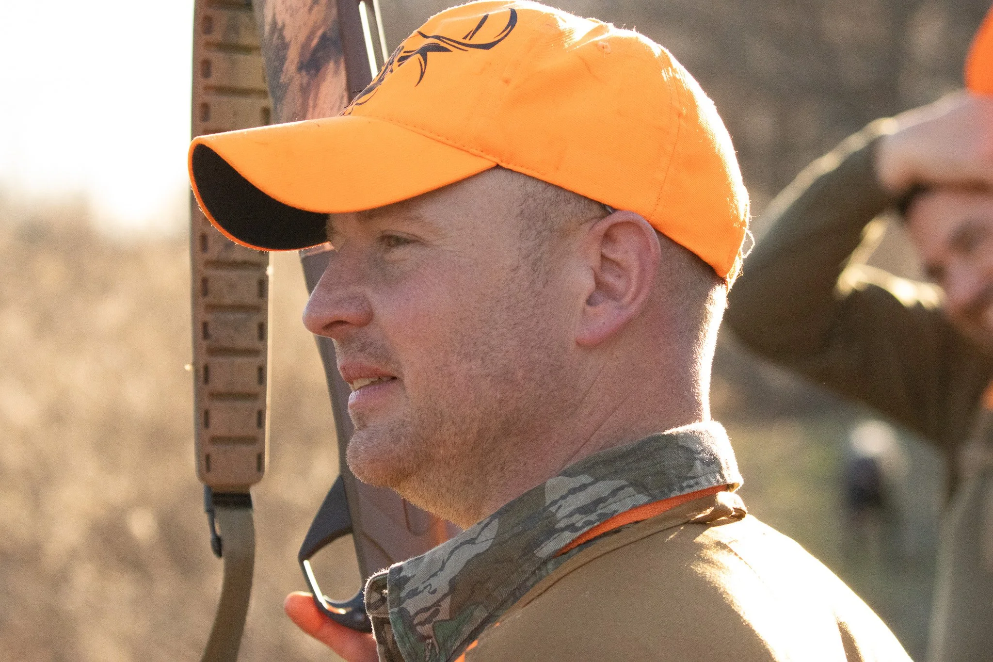 A soldier with short hair wearing an orange cap and camouflage clothing, holding a firearm in a dry outdoor setting.
