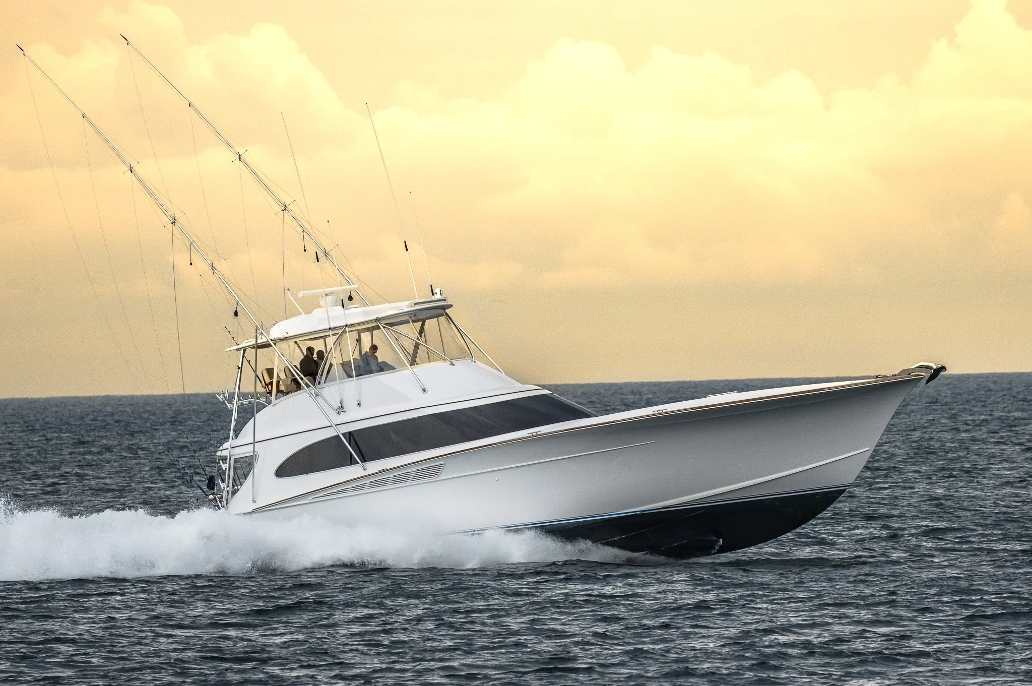 A white luxury yacht speeding across the water with three people visible on the deck, under a partly cloudy sky during daytime.