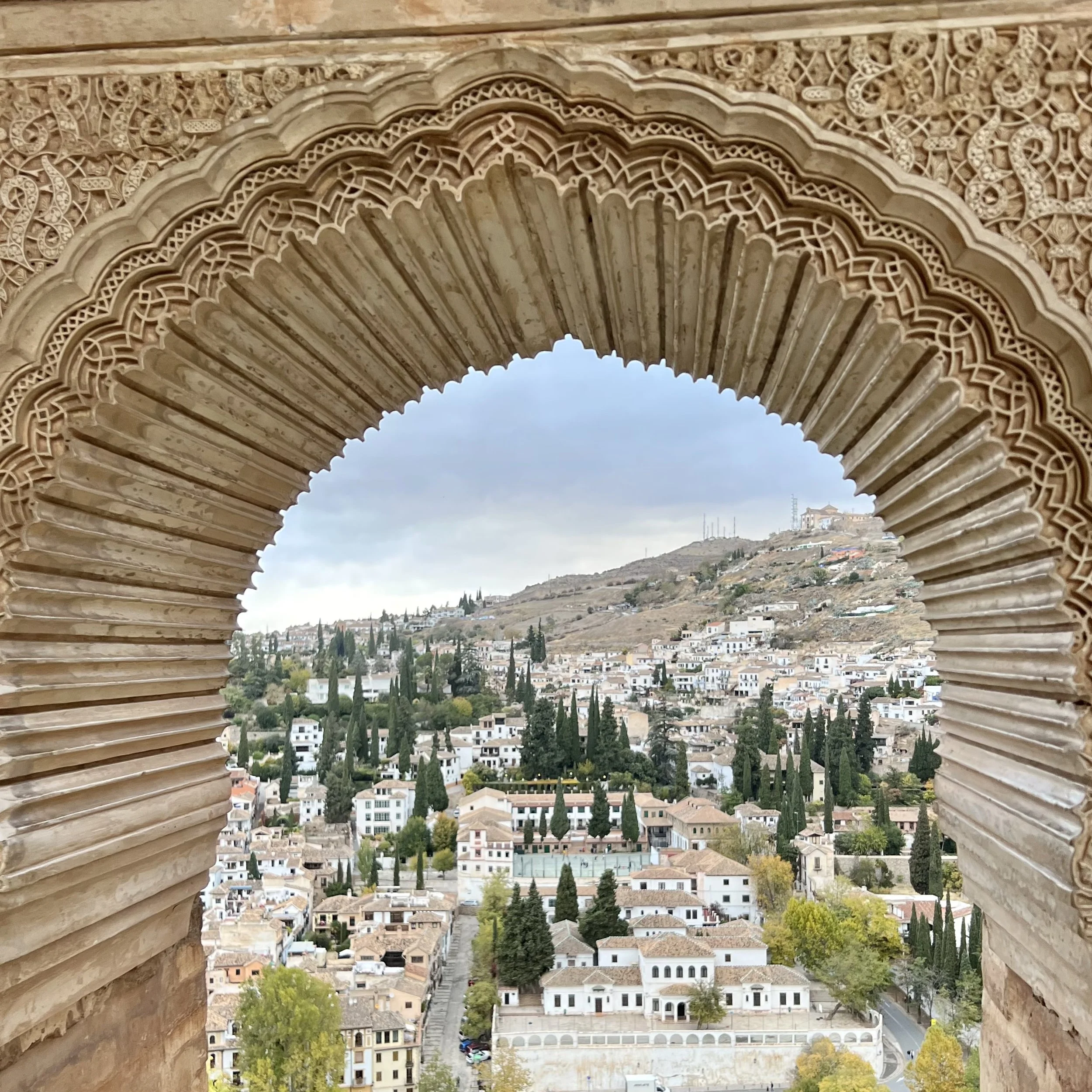 View through an ornate stone arch, showing a hillside with houses, trees, and cloudy sky.