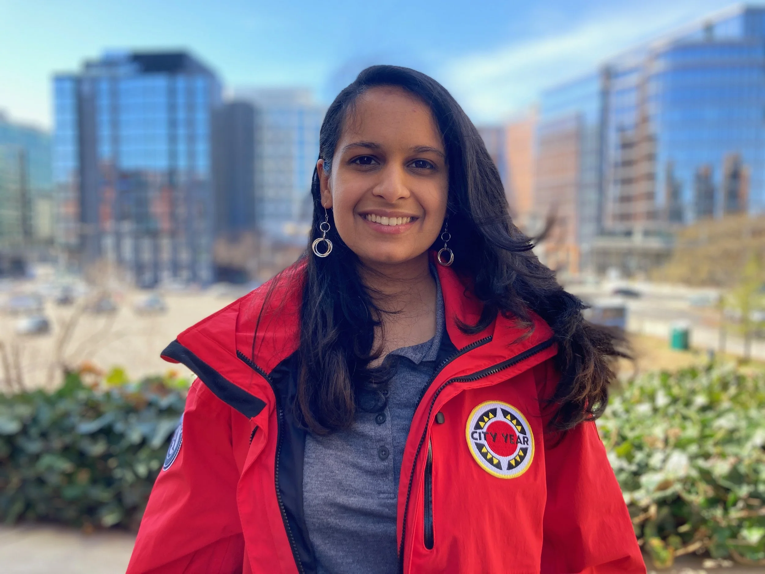 A woman with long dark hair wearing earrings and a red jacket with a City Year logo, standing outdoors with tall buildings in the background.