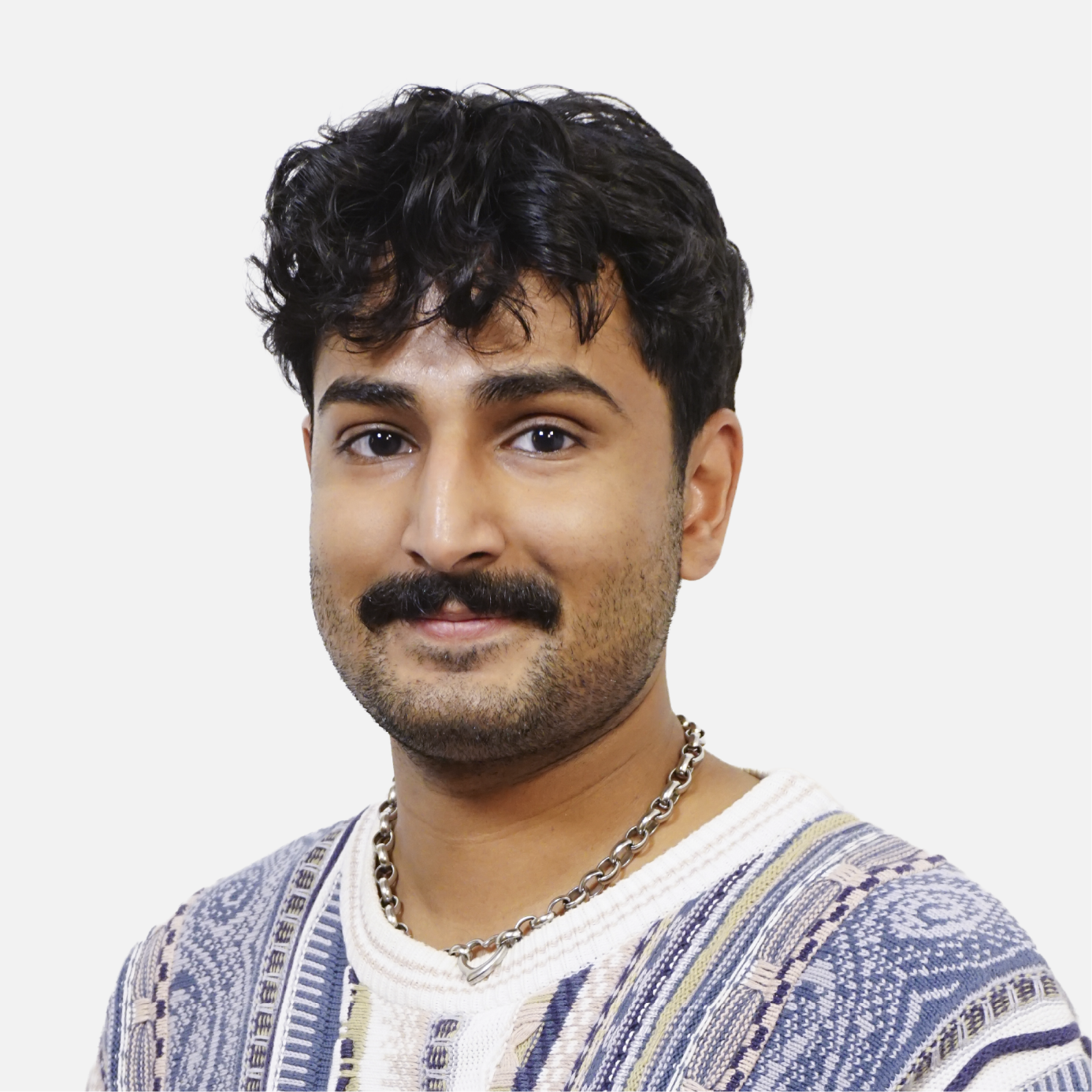 A young man with dark, curly hair, a mustache, and a beard, wearing a patterned sweater and a silver chain necklace, smiling at the camera against a plain white background.