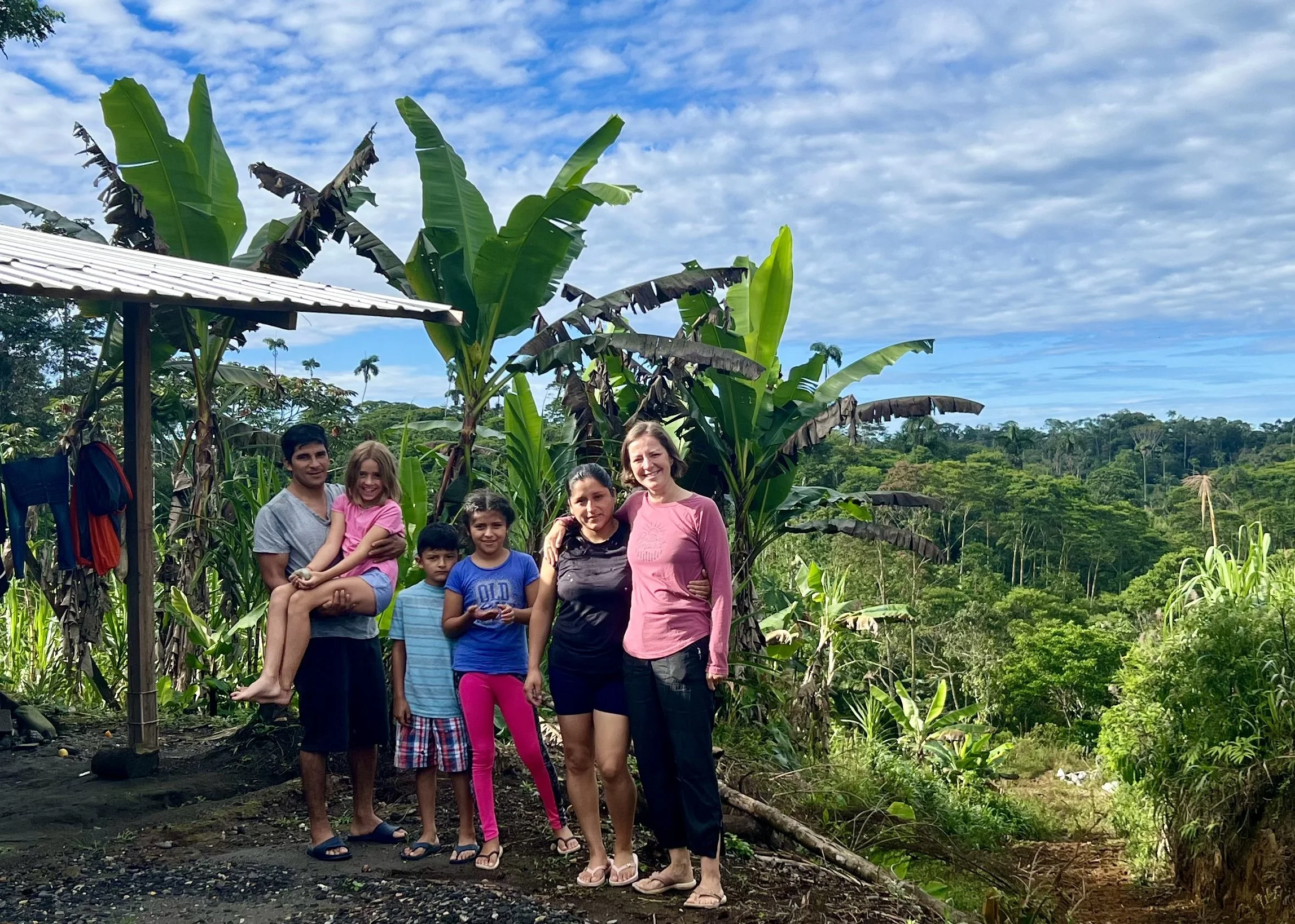 Group of people standing outdoors in a lush, tropical setting with banana plants and palm trees, under a partly cloudy sky.