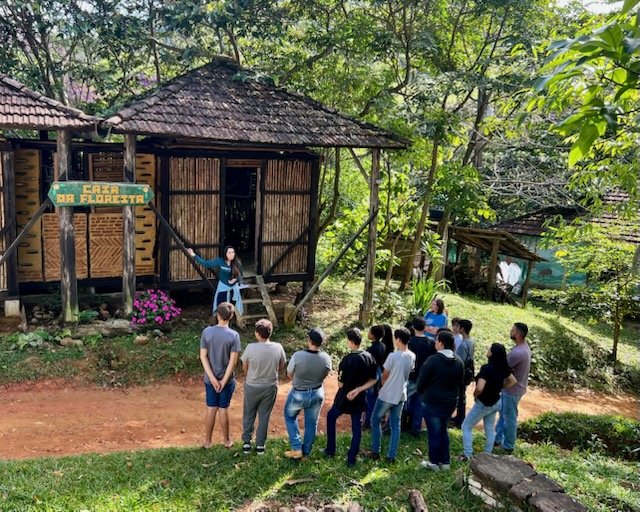 Group of people standing outside near a small wooden building in a lush, green forest setting, listening to a woman speaking.