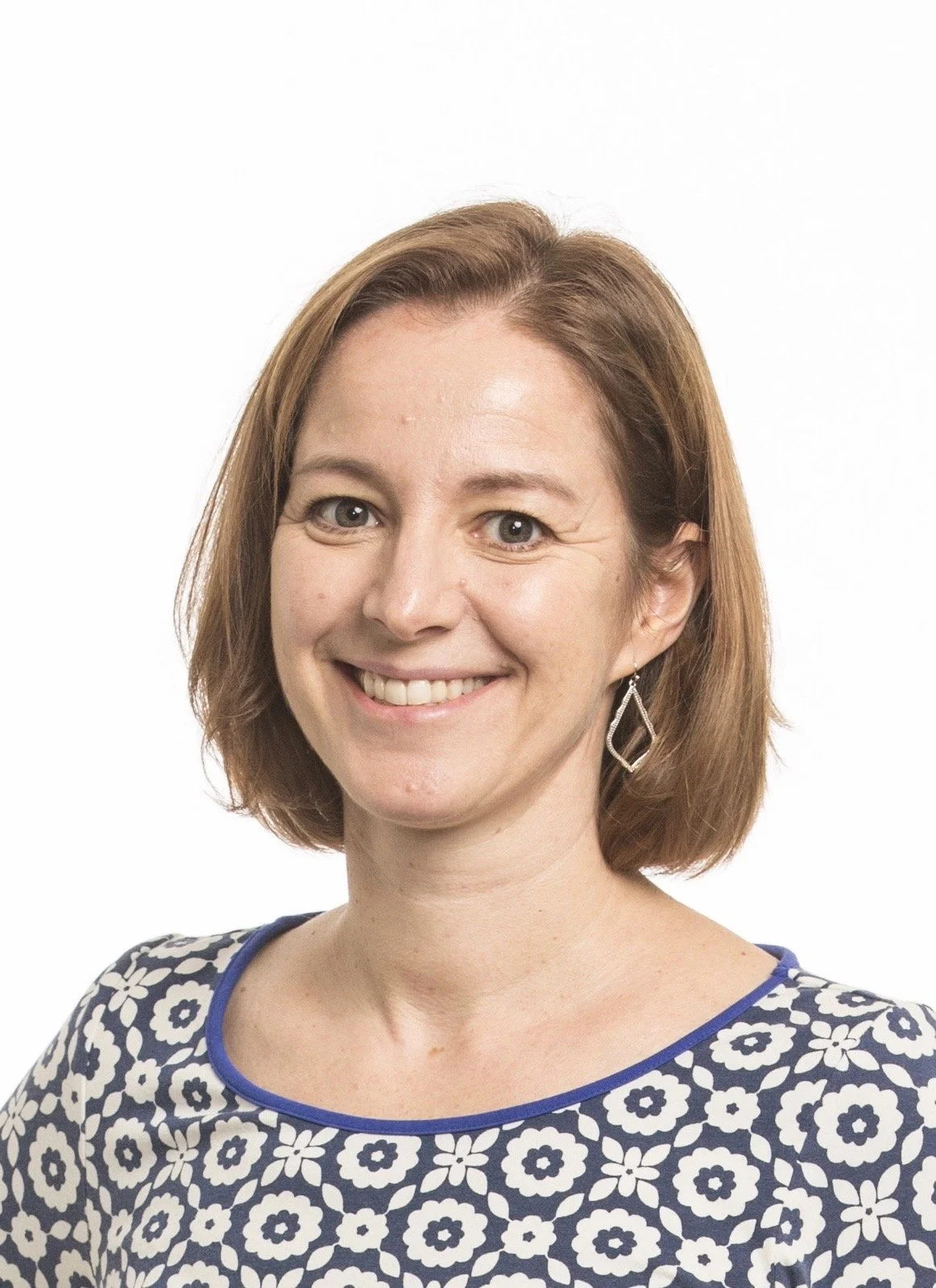 A woman with shoulder-length brown hair smiling against a white background, wearing a navy and white patterned top and silver earrings.