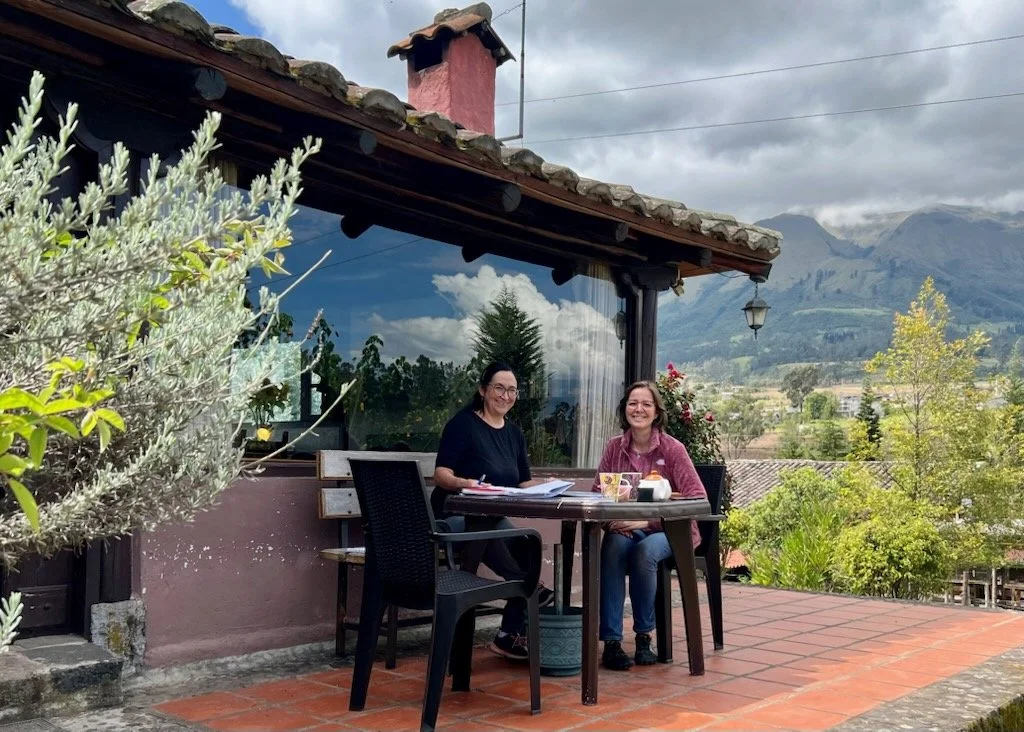 Two women sitting at a table on a patio with mountain scenery in the background.