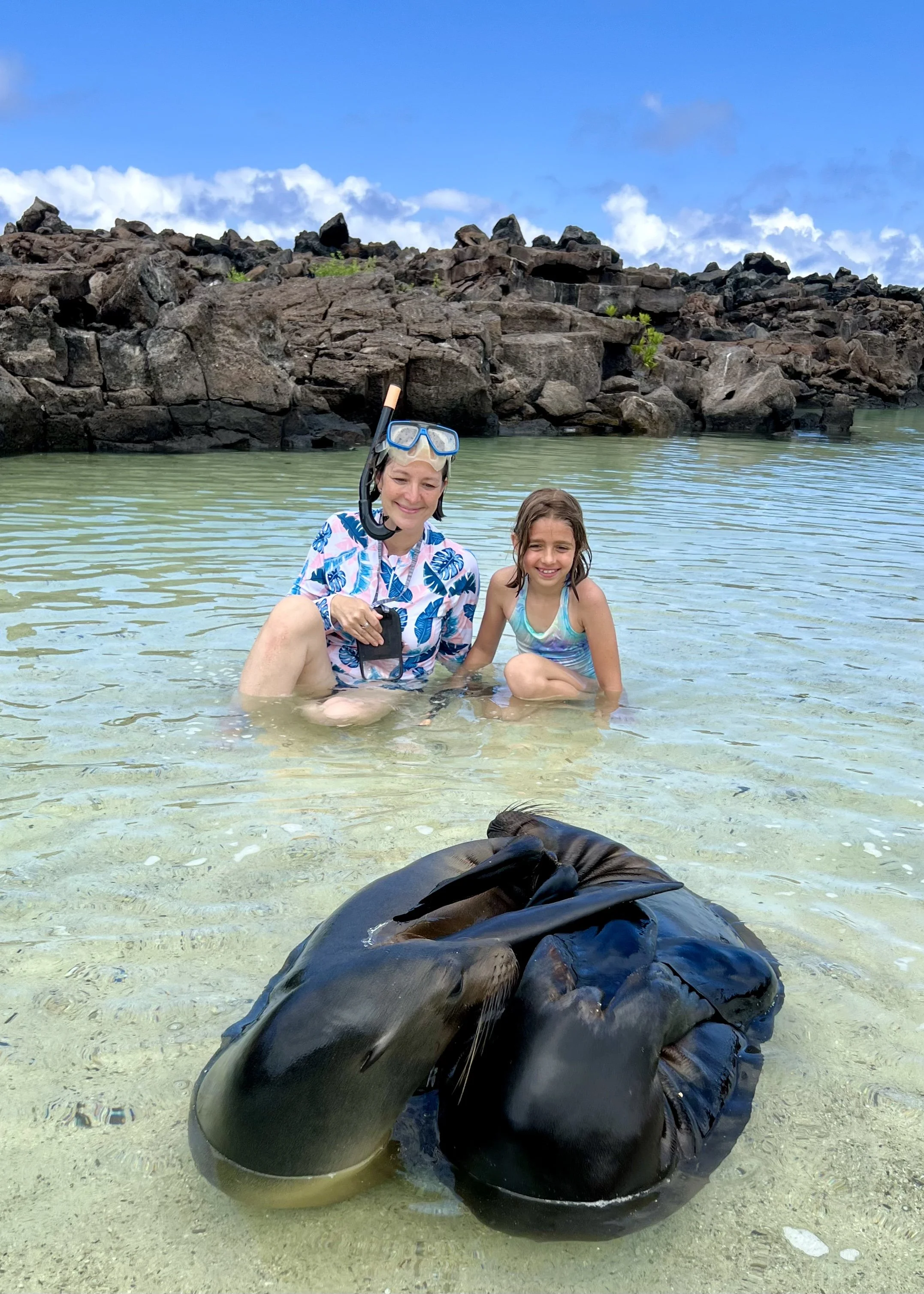 Two people, an adult woman and a young girl, sitting in shallow water on a beach or shoreline with a rocky formation and blue sky with clouds in the background. The woman is wearing a snorkel mask around her neck and patterned swimwear, and the girl is smiling in a colorful swimsuit. A large, dark, smooth object, possibly a marine animal or a piece of debris, is in the water in the foreground.