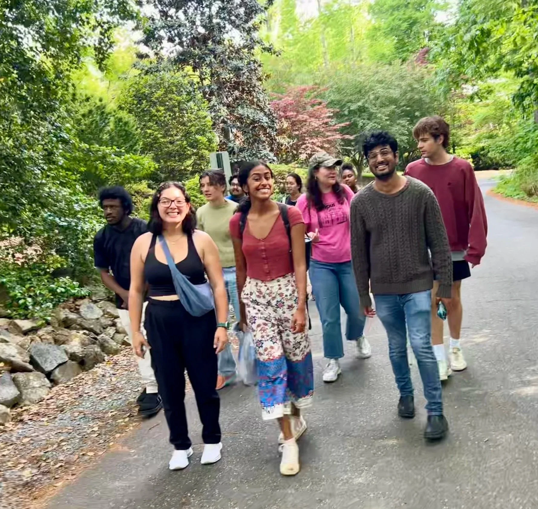 A diverse group of young adults walking along a paved trail surrounded by lush green trees. They are smiling and appear to be enjoying a hike or walk outdoor activity.