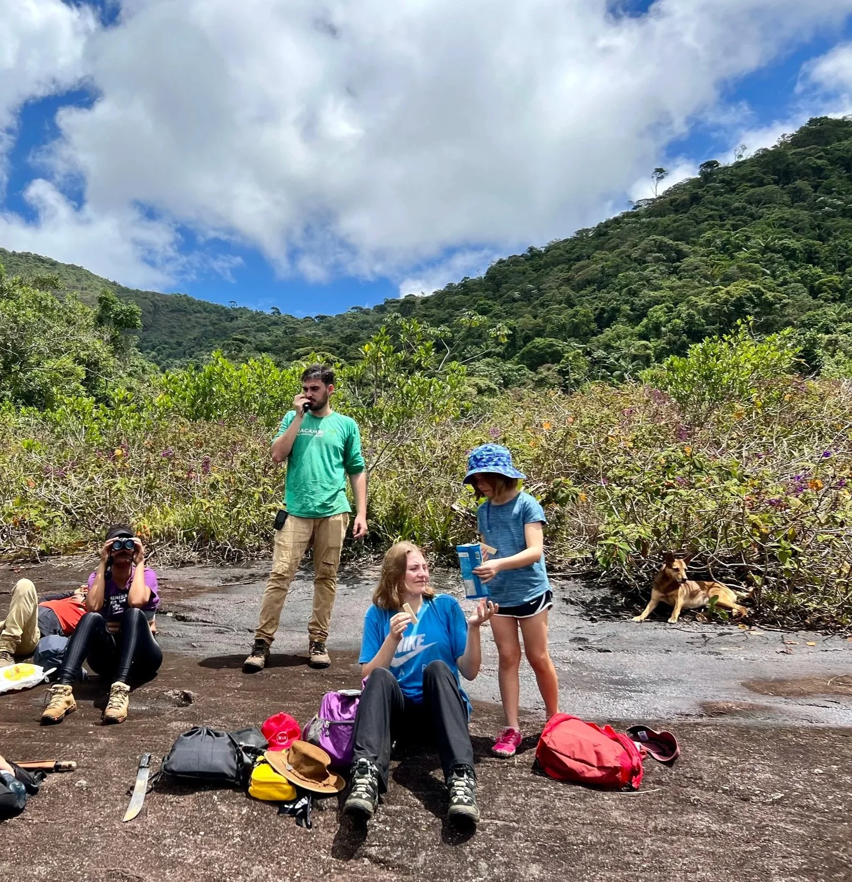 Group of people resting on a volcanic ash landscape surrounded by lush green hills and a partly cloudy blue sky. Gear such as backpacks, shoes, and binoculars are on the ground.