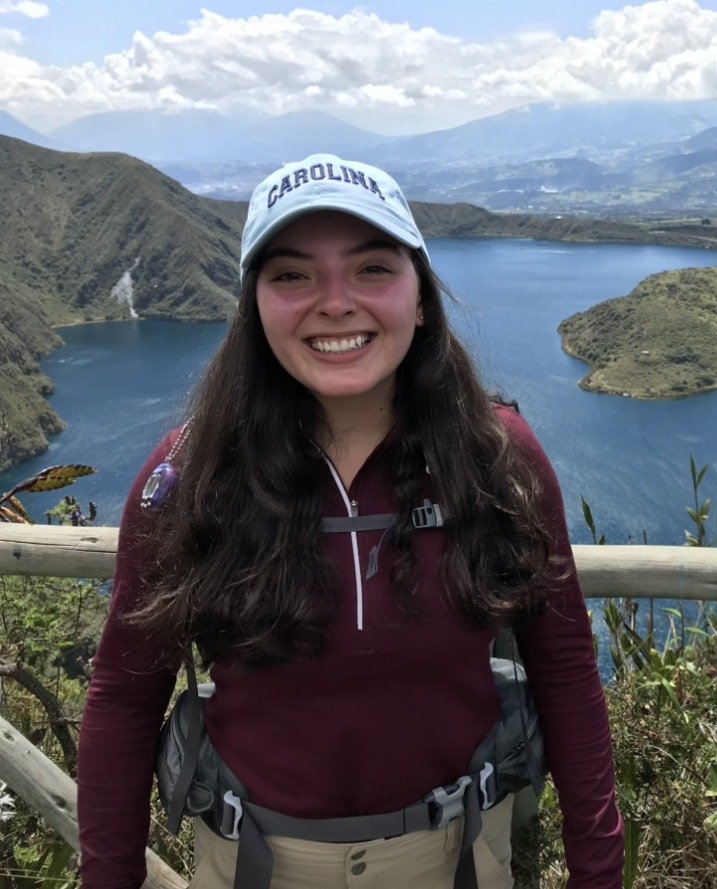 Young woman smiling outdoors near a wooden fence, with a scenic lake and mountainous landscape in the background.