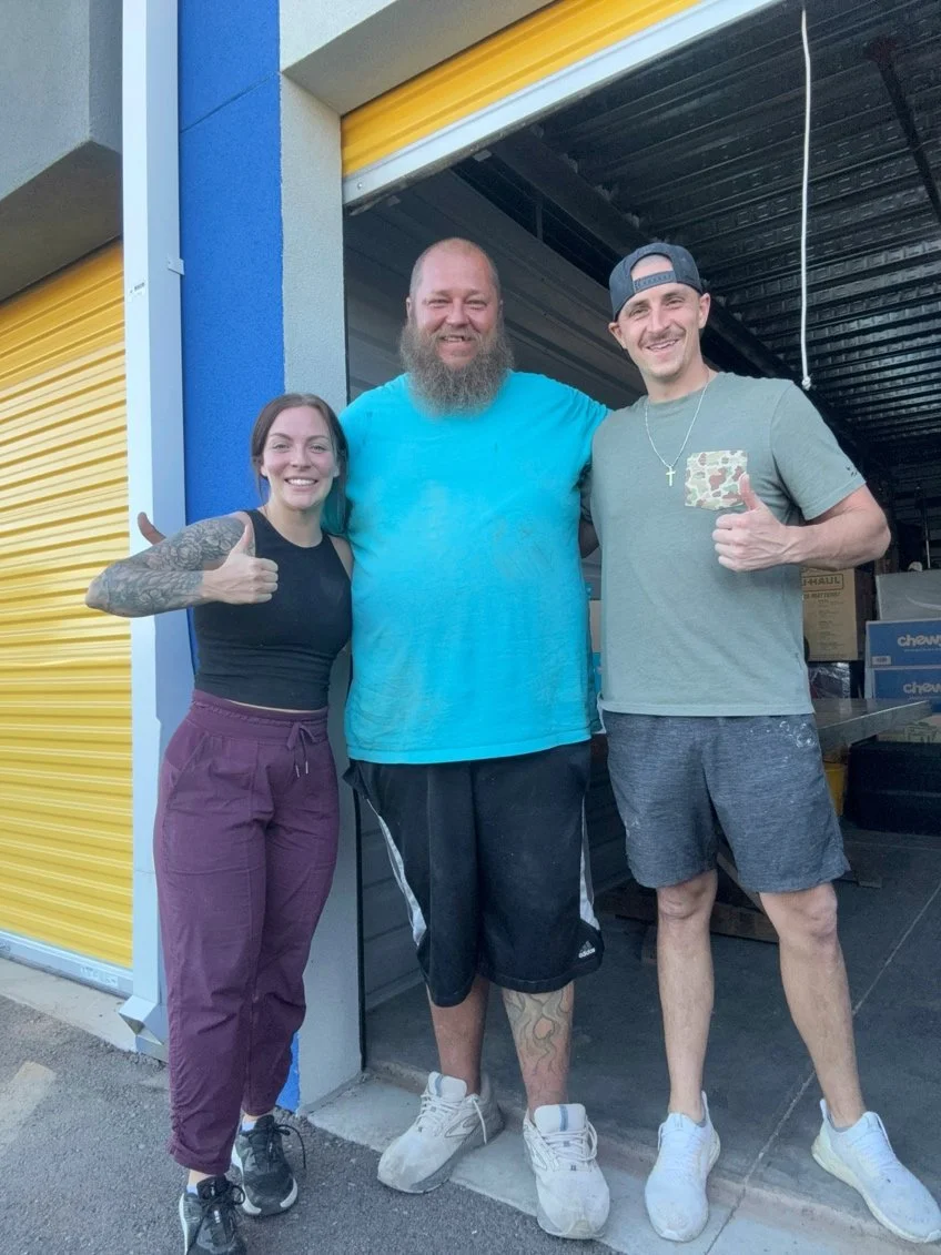 Three people standing outside a storage unit, smiling and giving thumbs up. The person on the left has tattoos on her right arm, the person in the middle has a beard, and the person on the right is wearing a gray cap backward.