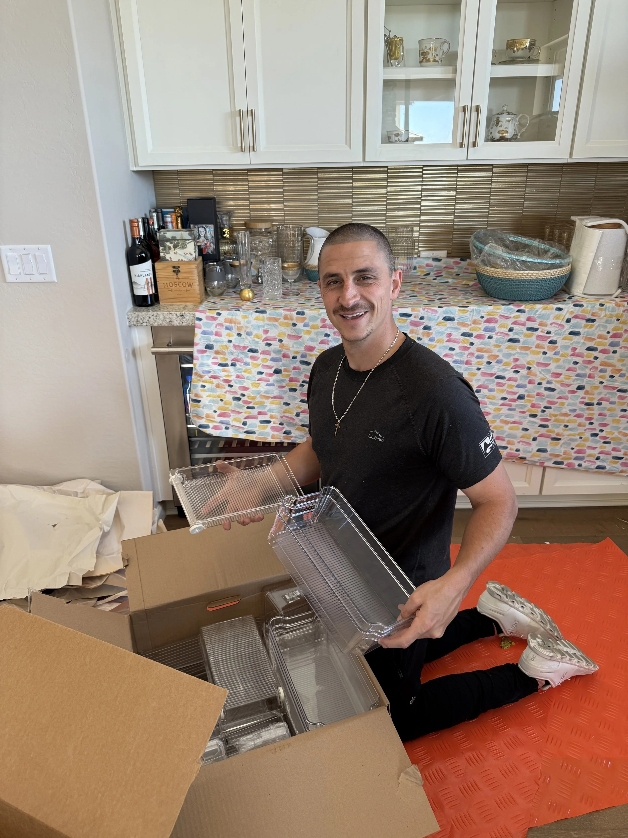 A man with short hair and a cross necklace kneeling on an orange mat, opening a clear plastic container in front of a large cardboard box. The background shows a kitchen counter with wine bottles, glassware, and decorative items, and white cabinets with glass doors.