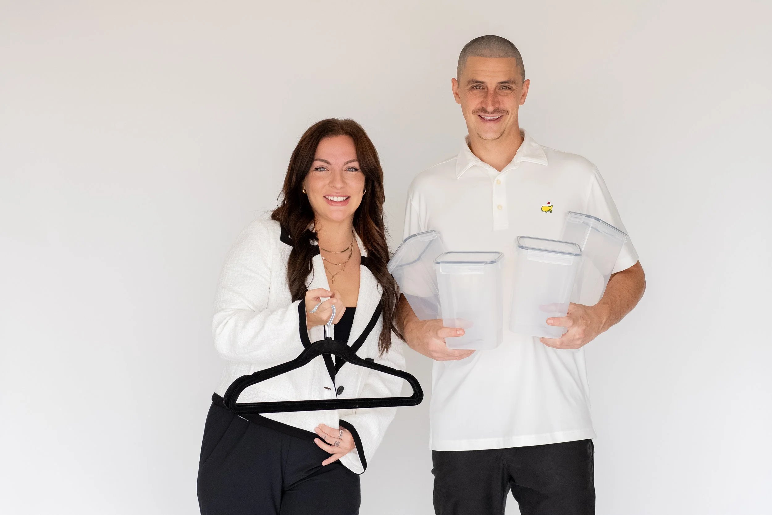 A woman and a man smiling, standing side by side against a plain white background, holding empty plastic containers and a black hanger.
