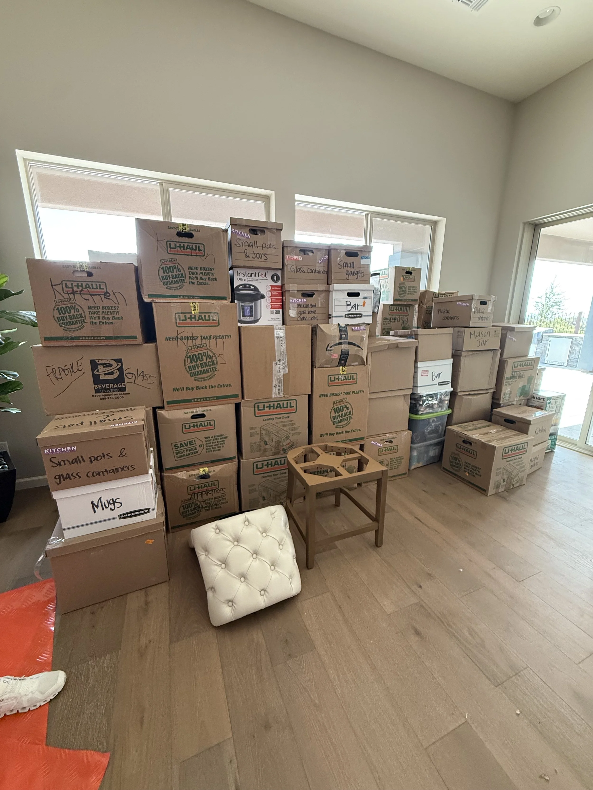 An interior room with stacked boxes labeled for various storage items, a small padded stool, and a wooden plant stand with a plant. Large windows and a sliding glass door are visible, allowing natural light to enter.