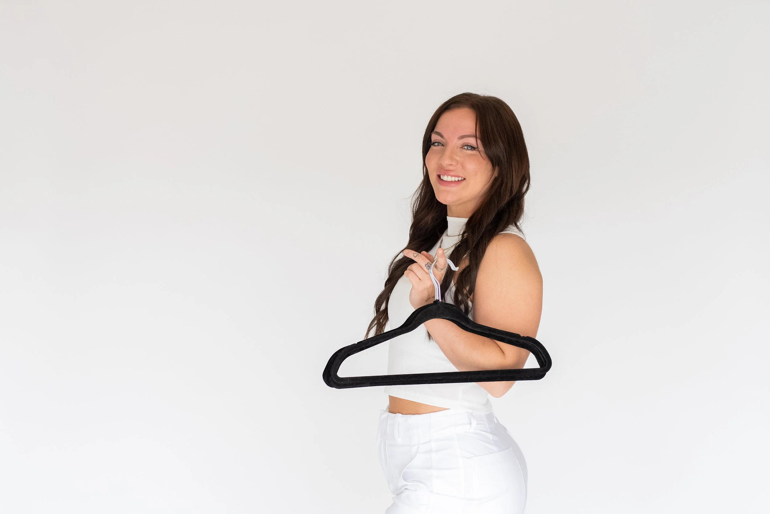A young woman with long brown hair and a white top is smiling and holding a black clothes hanger against a plain white background.