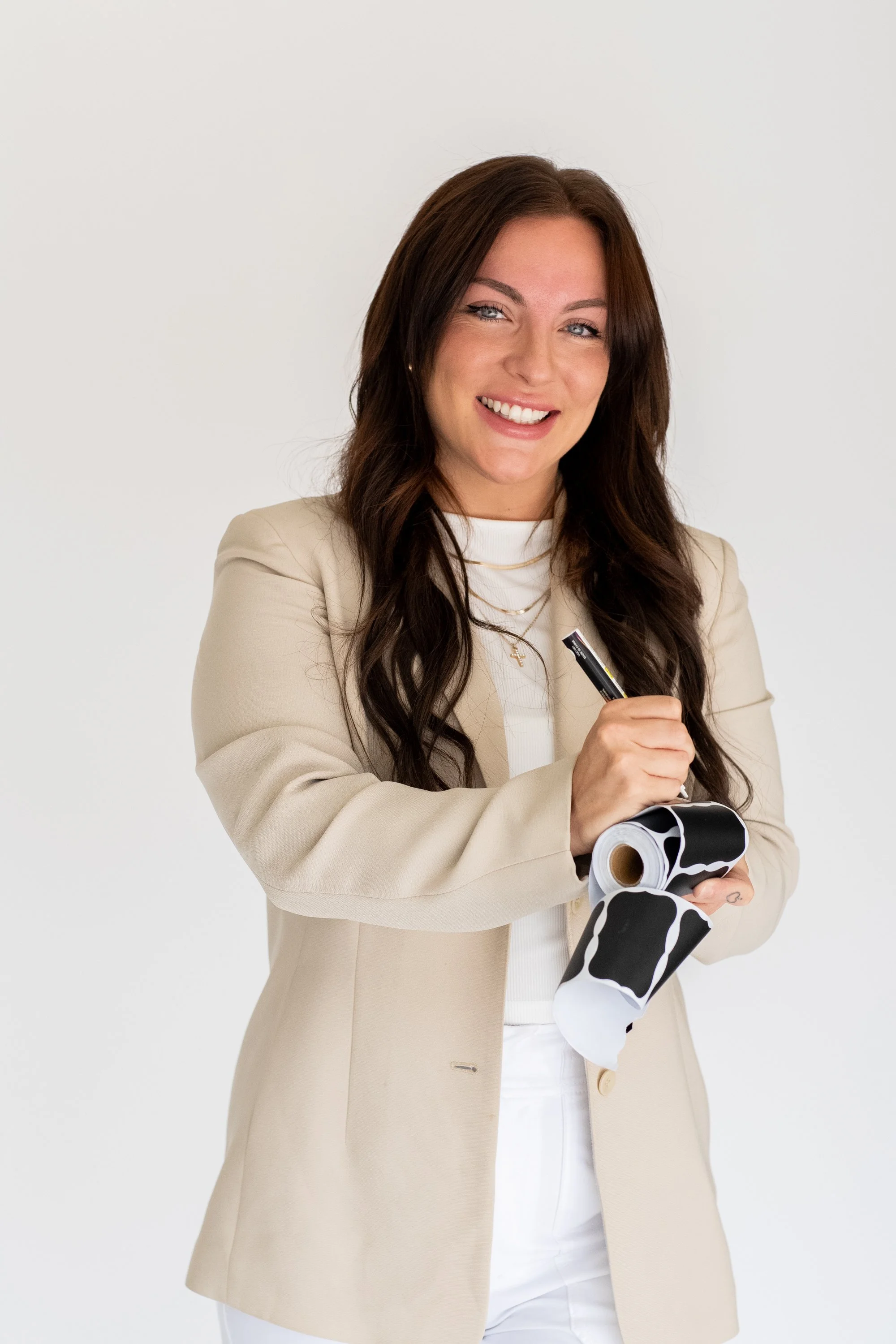 A woman with long brown hair, wearing a beige blazer, white top, and white pants, smiling and holding a marker and a roll of wallpaper with a black and white animal print pattern.