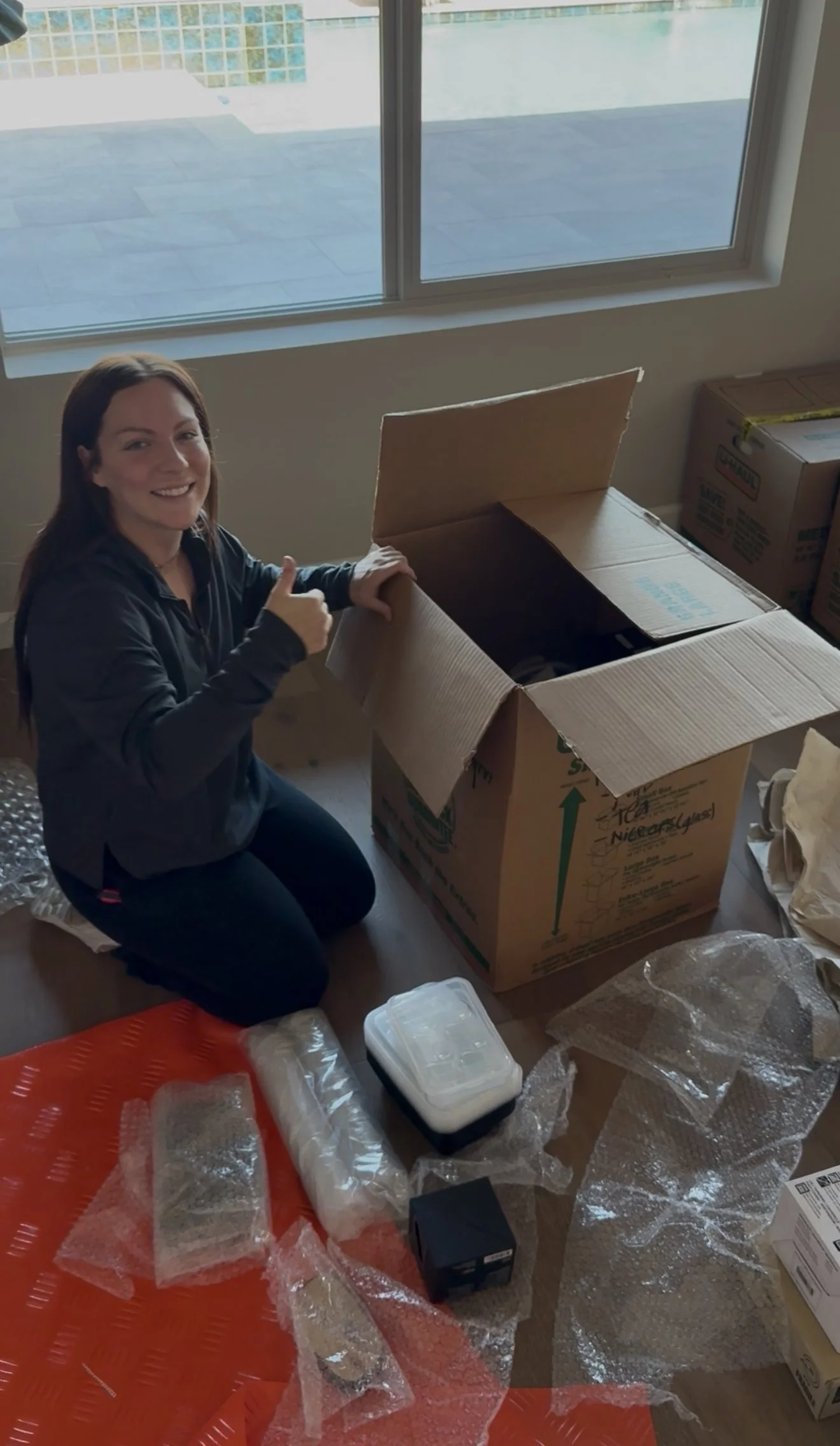 A woman with long brown hair wearing a dark jacket kneels inside a room, giving a thumbs-up, next to a large open cardboard box. Items like plastic wrap, a small container, and various packaging materials are scattered on the floor. A window shows a view of an outdoor patio with a tiled surface.