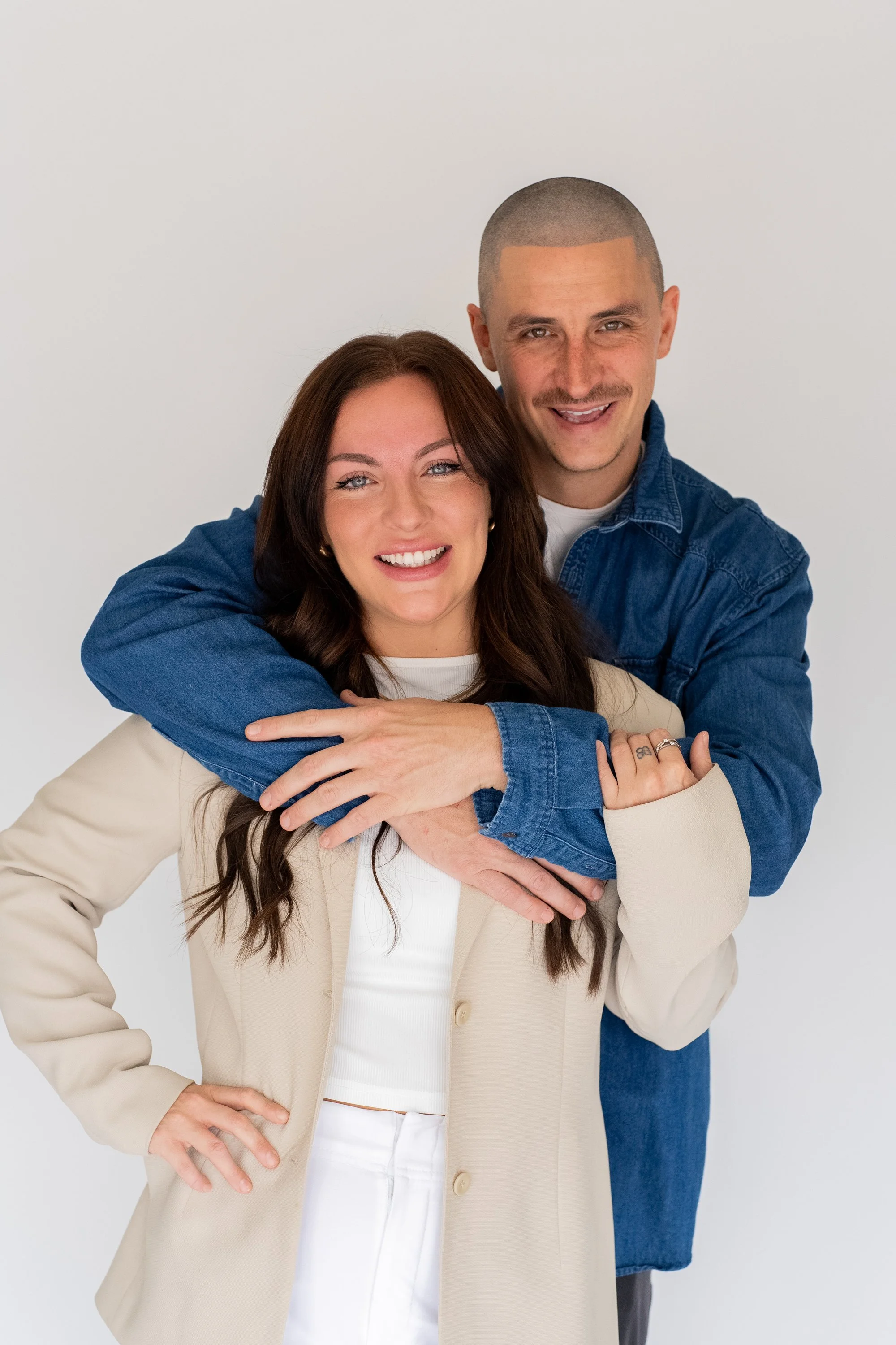 Two smiling individuals, a woman with long brown hair and a man with a shaved head, embrace against a plain white background. The woman is wearing a beige blazer and white top, and the man is in a blue denim jacket.
