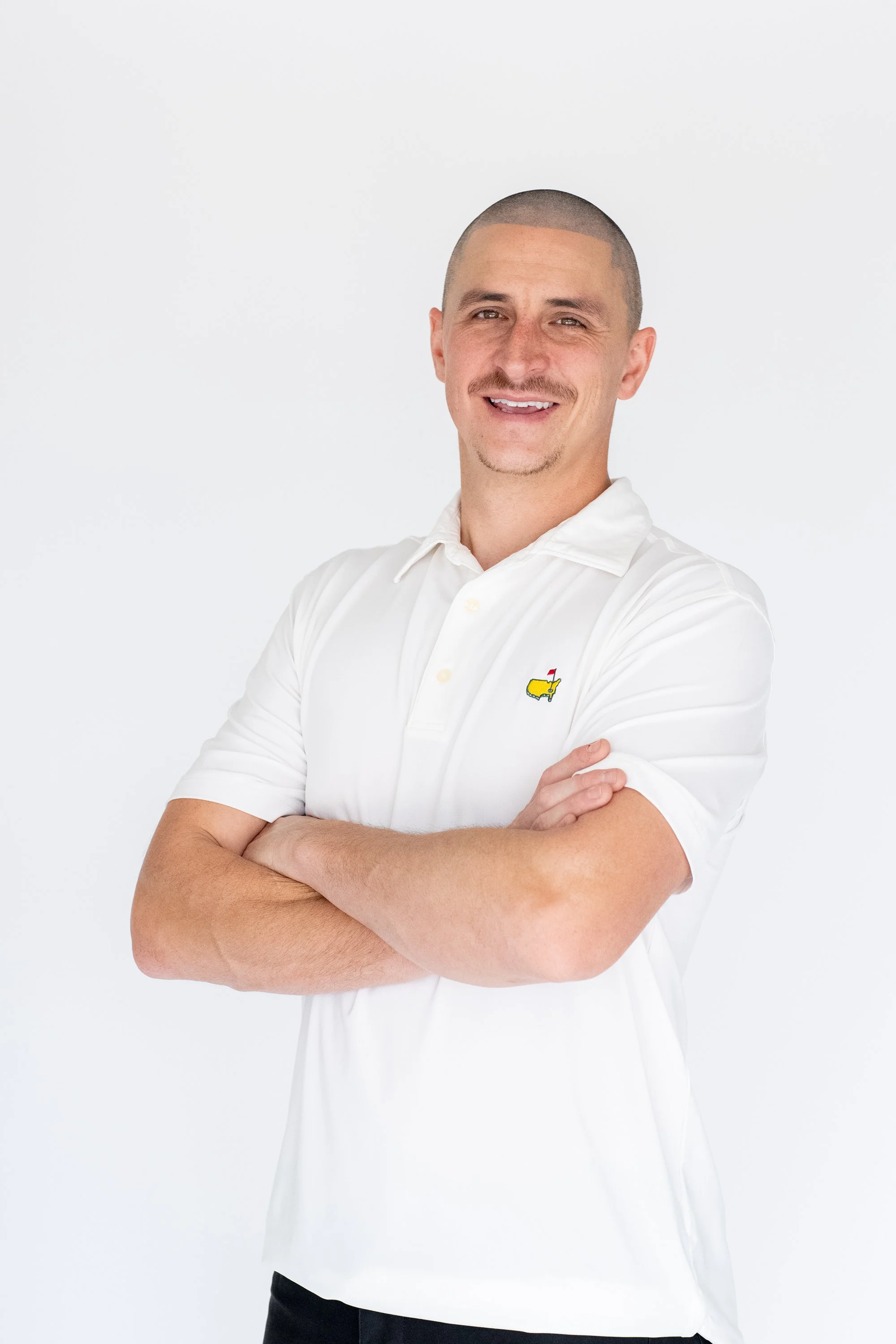 A man with a shaved head and a light goatee, wearing a white polo shirt with a small yellow logo of a dog on the chest, smiling with arms crossed in front of a plain white background.