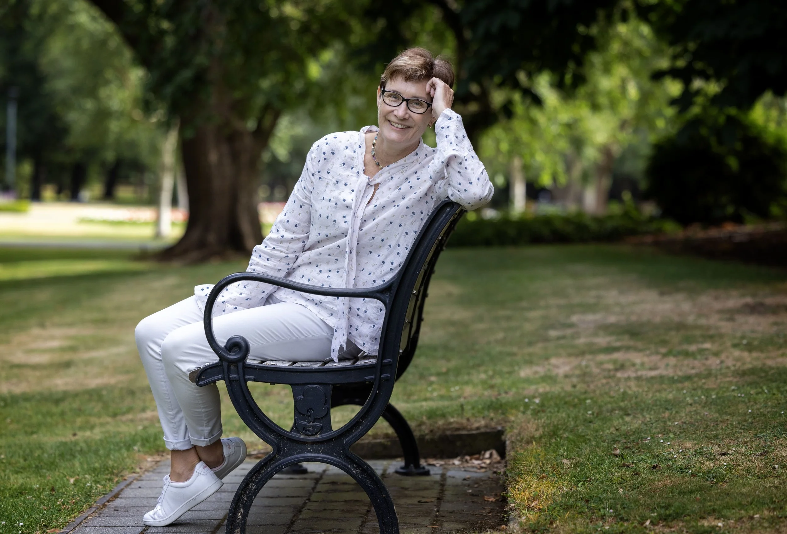 A woman sitting on a park bench, smiling, wearing glasses, a white blouse with blue polka dots, white pants, and white sneakers, in a lush outdoor setting with trees and grass.