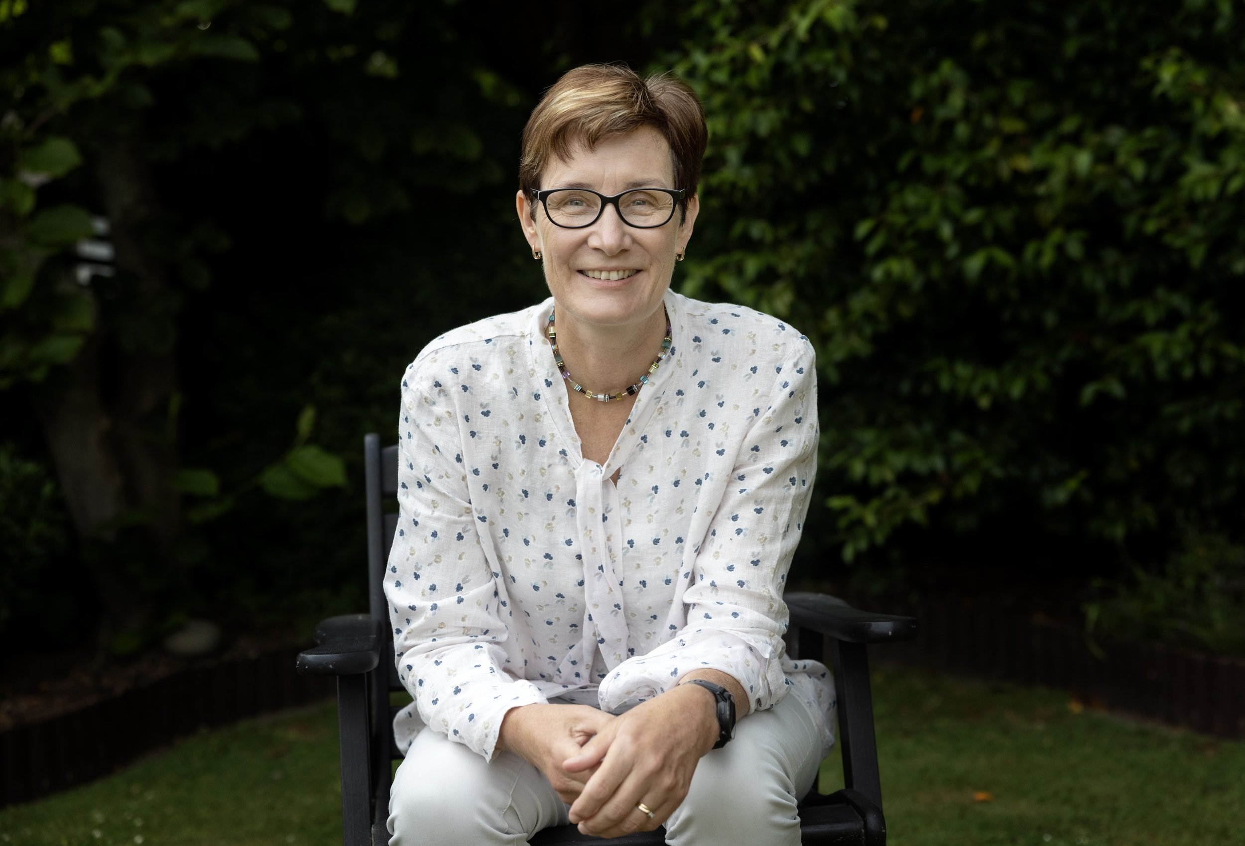 A woman with short brown hair and glasses, wearing a white blouse with small blue patterns, sitting on a black chair outdoors with green bushes in the background.