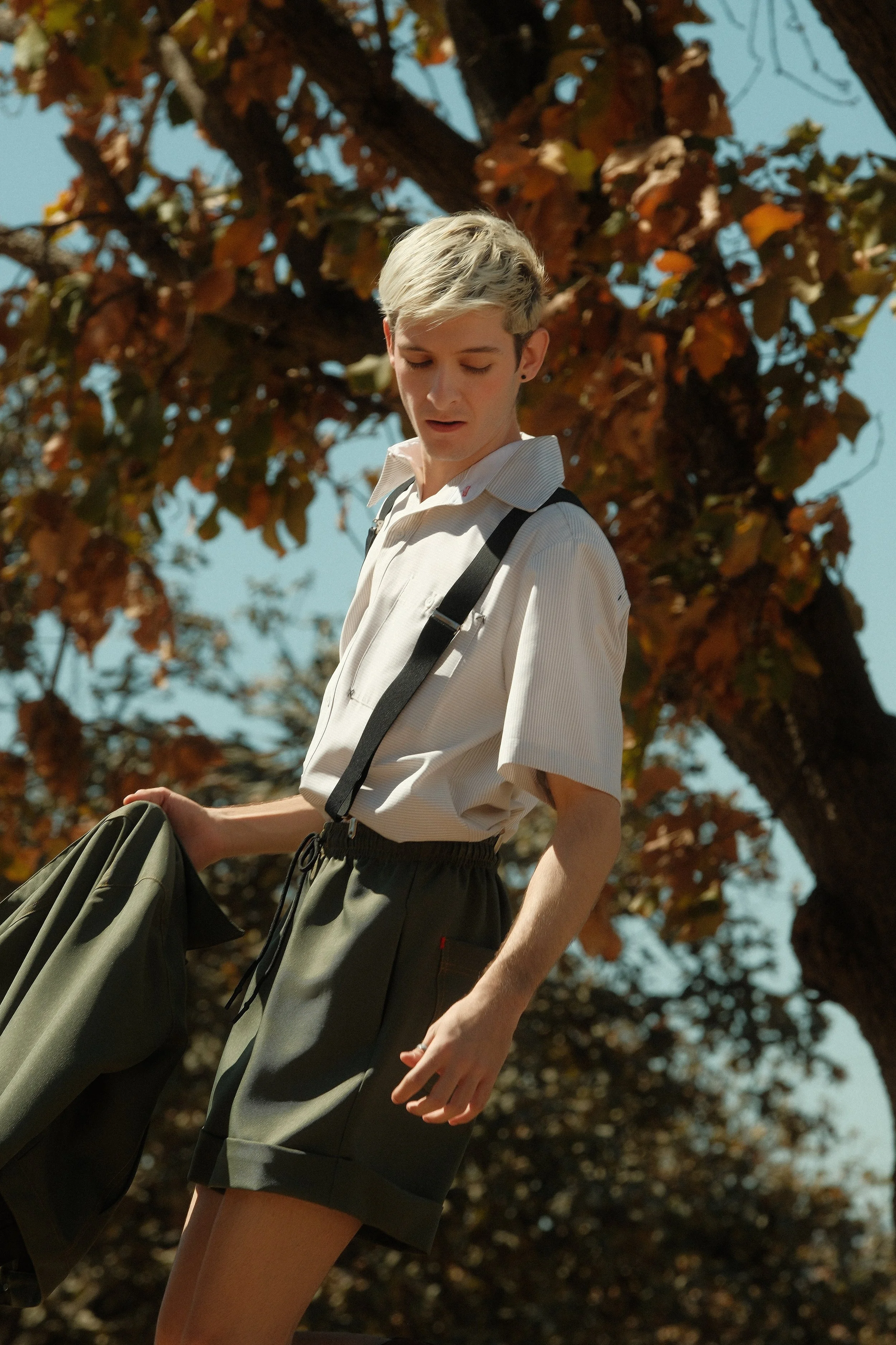 Joven con cabello rubio y peinado de lado, vistiendo una camisa blanca de manga corta, pantalones cortos oscuros, con una mochila y sosteniendo una chaqueta, de pie junto a un árbol con hojas otoñales y cielo despejado.