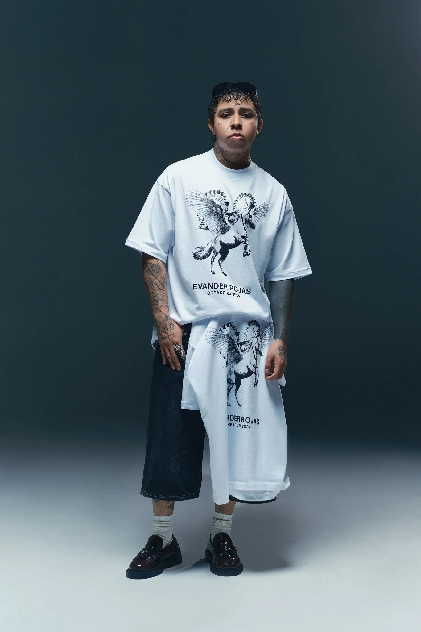 Persona con peinado rizado, vestida con camiseta blanca con un diseño de grifo y una pegatina de un unicornio con alas, shorts de mezclilla y accesorios como anillos y tatuajes en brazos y cuello, de pie contra fondo oscuro y claro.