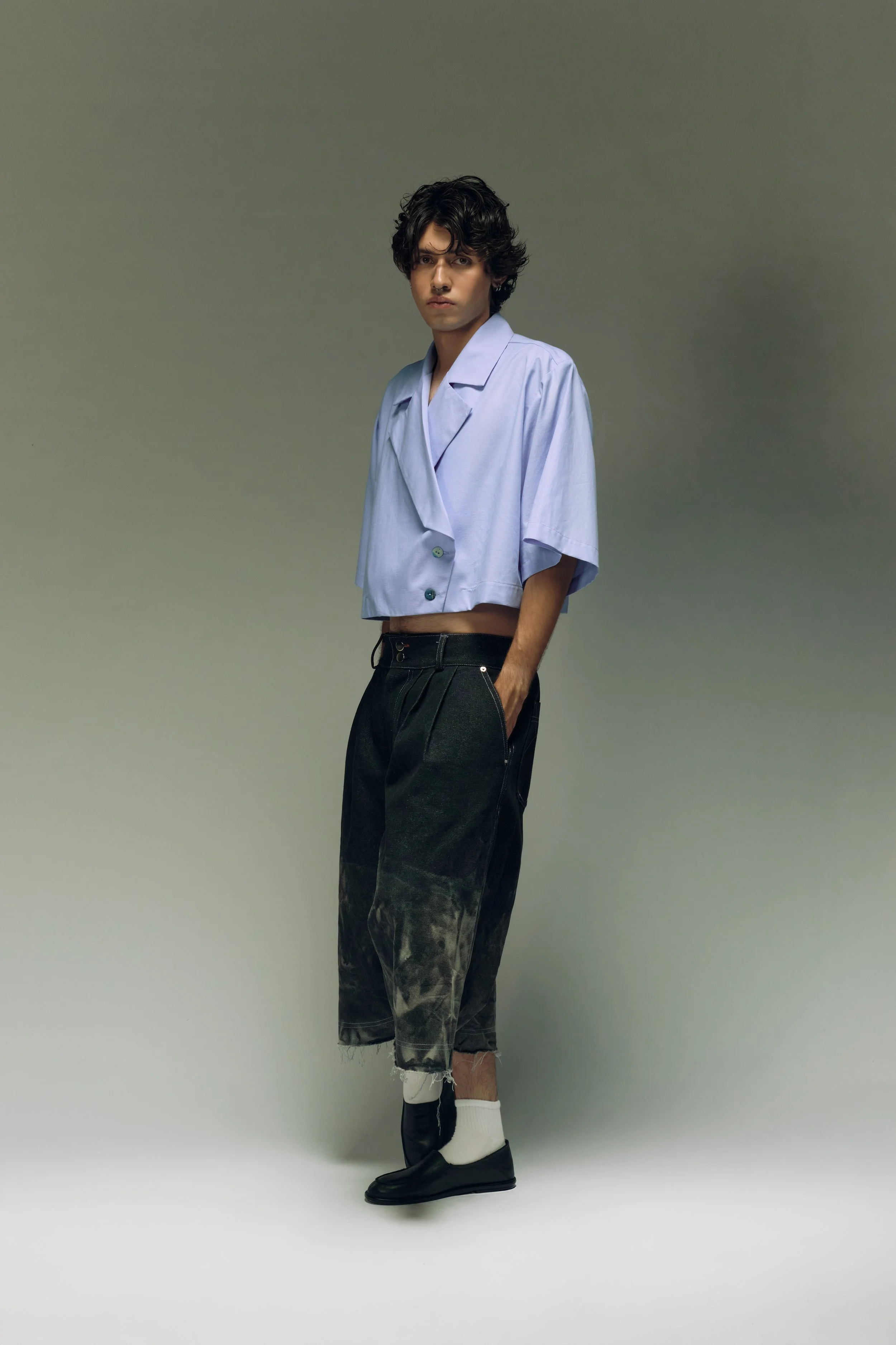 Joven con cabello rizado, camiseta de pijama azul, pantalones negros con estilo desgastado y zapatos negros, en fondo gris.