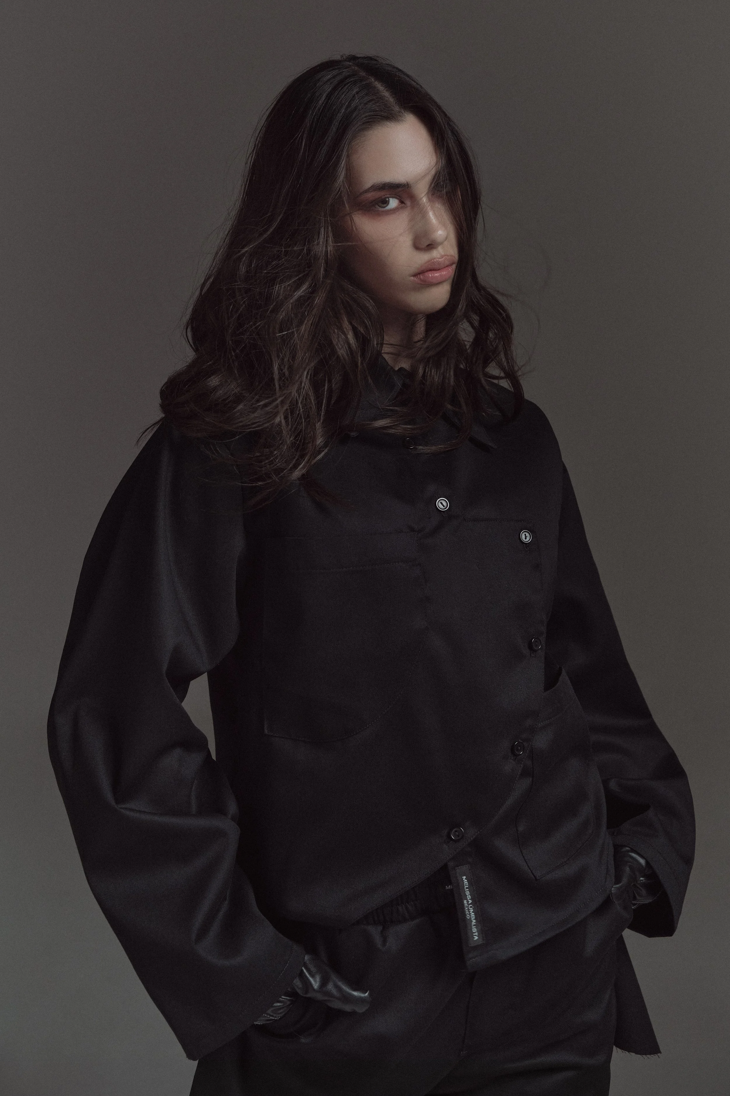 Mujer con cabello oscuro y ojeras, vestida con chaqueta negra, en un fondo gris, con expresión seria y postura relajada.