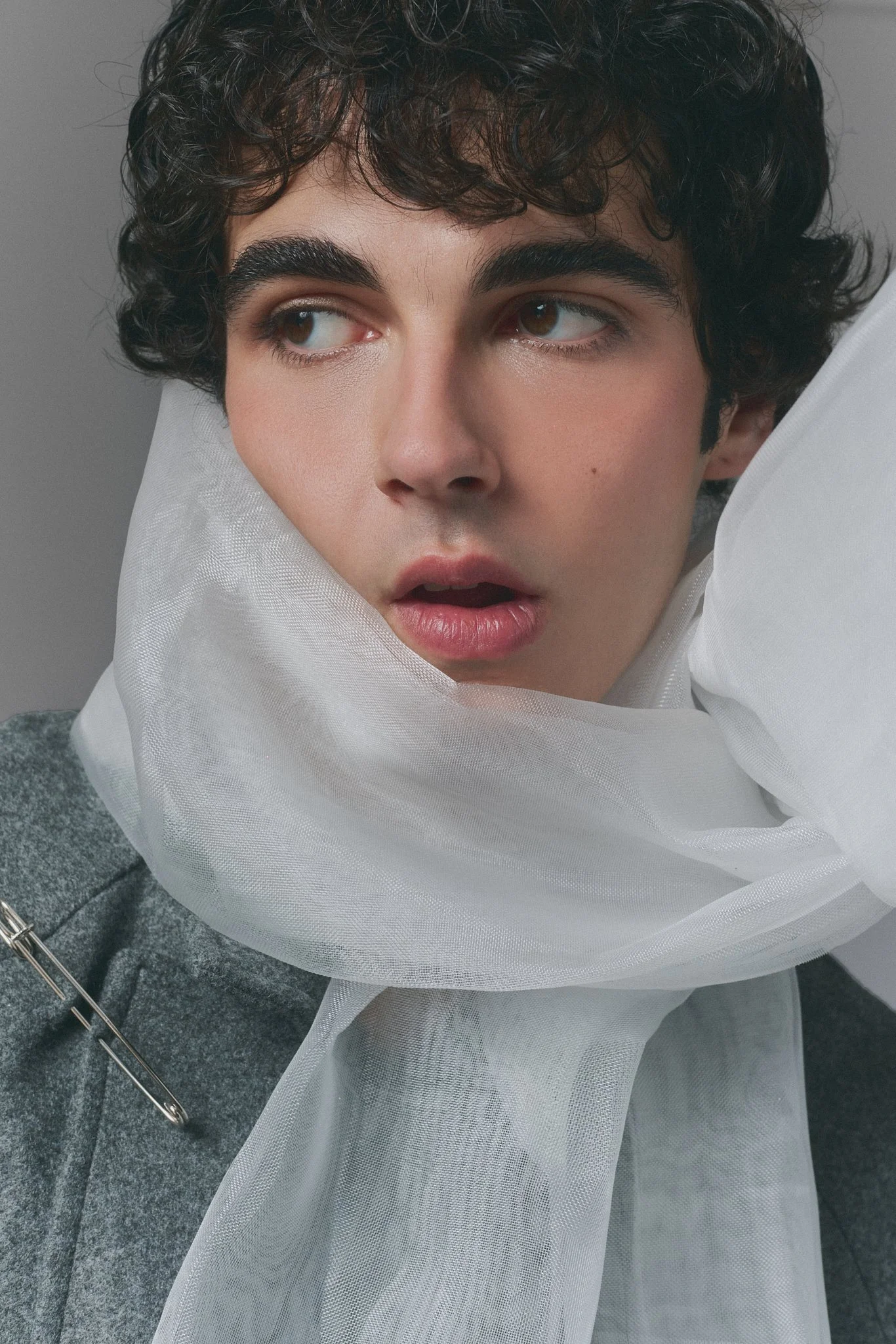 Primer plano de un rostro de joven con cabello rizado rubio y piel clara, rodeado de tela blanca y fondo gris. La expresión facial es sorprendida o pensativa.