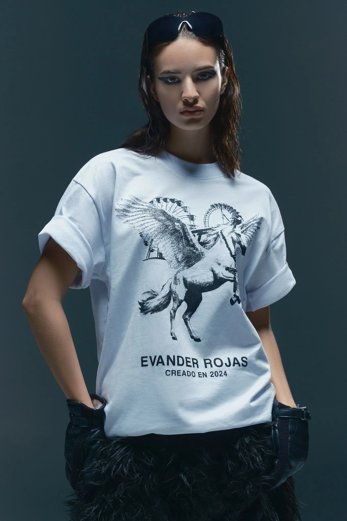 Joven mujer con gafas en la cabeza, con camiseta blanca con dibujo de un unicornio y una noria, y pantalones con peluche negro, de cabello castaño y piel clara, frente a fondo gris oscuro.