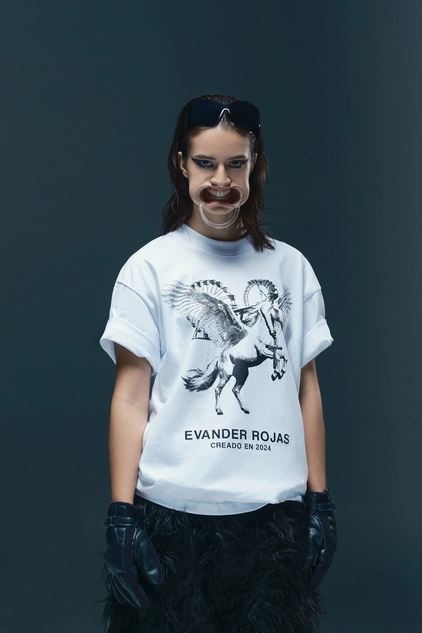 Una joven con cabello oscuro y lentes de sol en la cabeza lleva una camiseta blanca con un diseño de un unicornio y texto que dice 'Evander Rojas, Creado en 2024'. Ella lleva guantes negros y pantalones negros con un diseño de plumas.